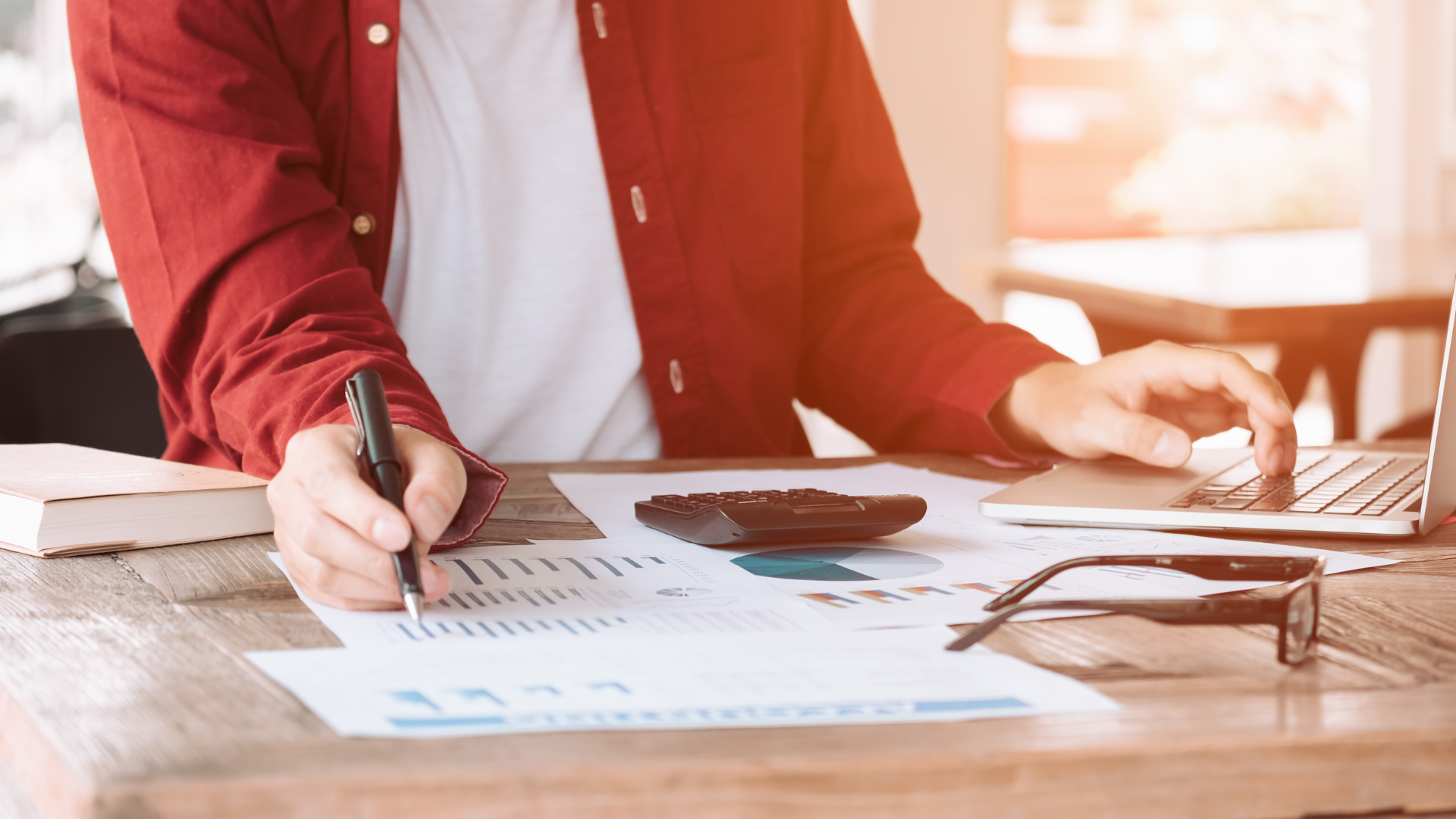 Person in red shirt working on finances with laptop, calculator, and papers.