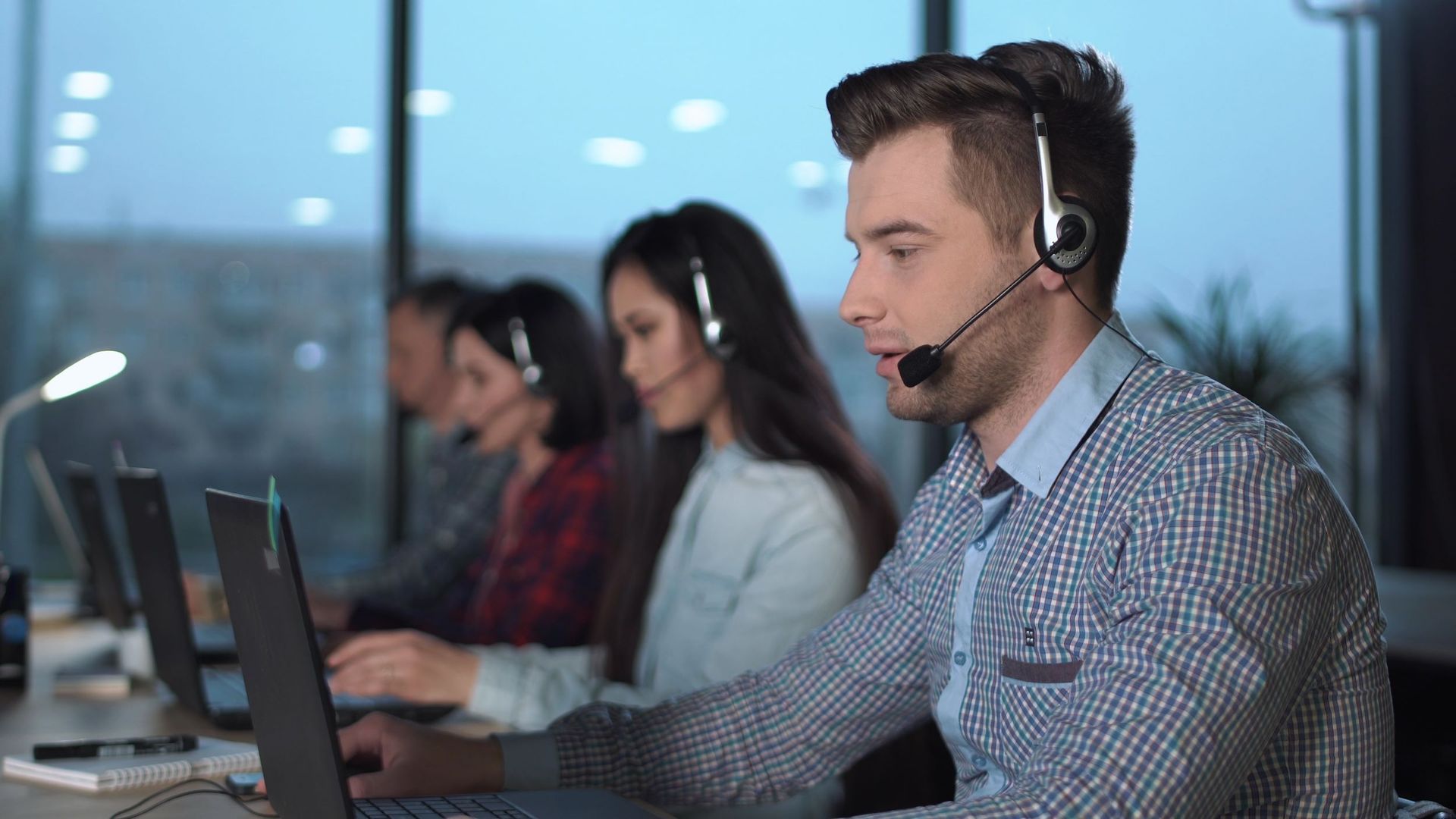 Call center employees wearing headsets, working on laptops in a dimly lit office.