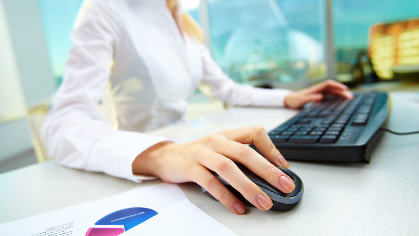 Woman at desk, hands on mouse and keyboard, looking at a computer screen.