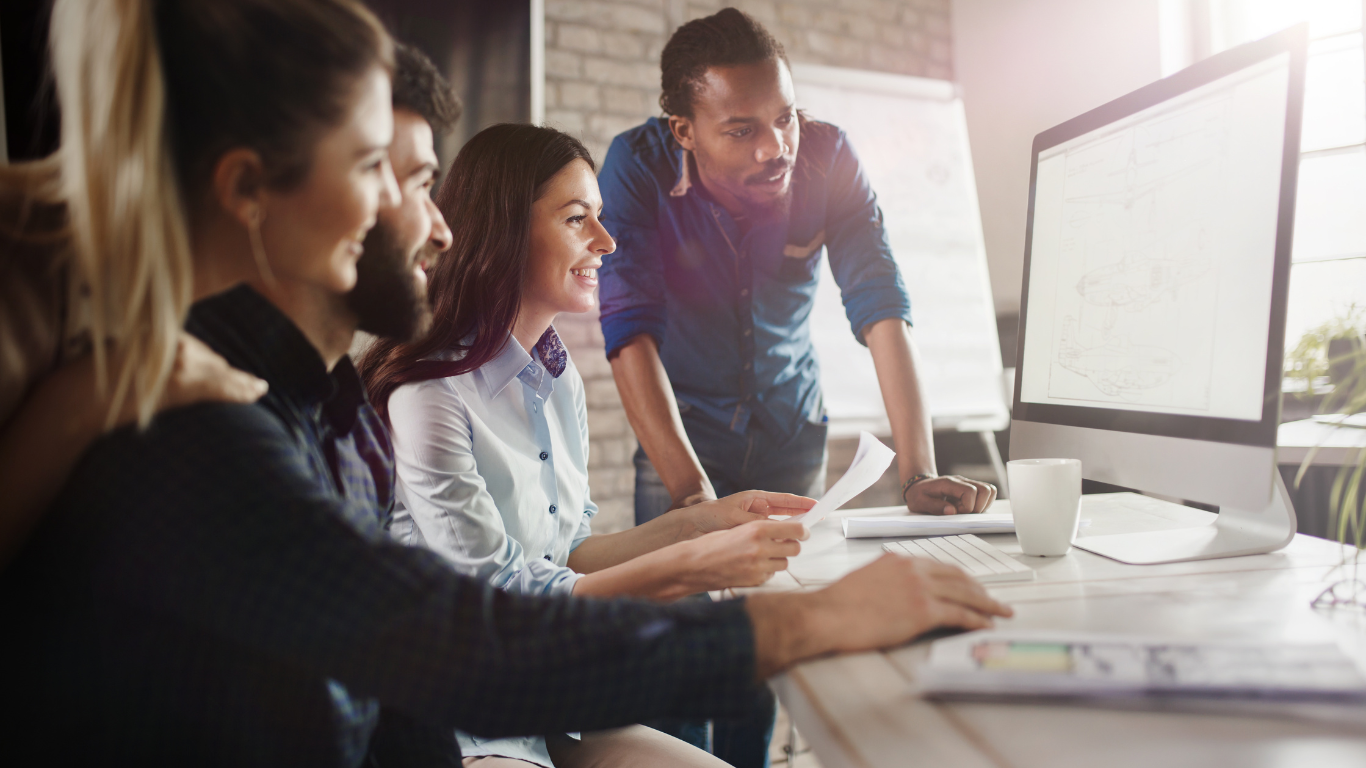 Group of people at desk looking at computer screen, discussing paperwork.