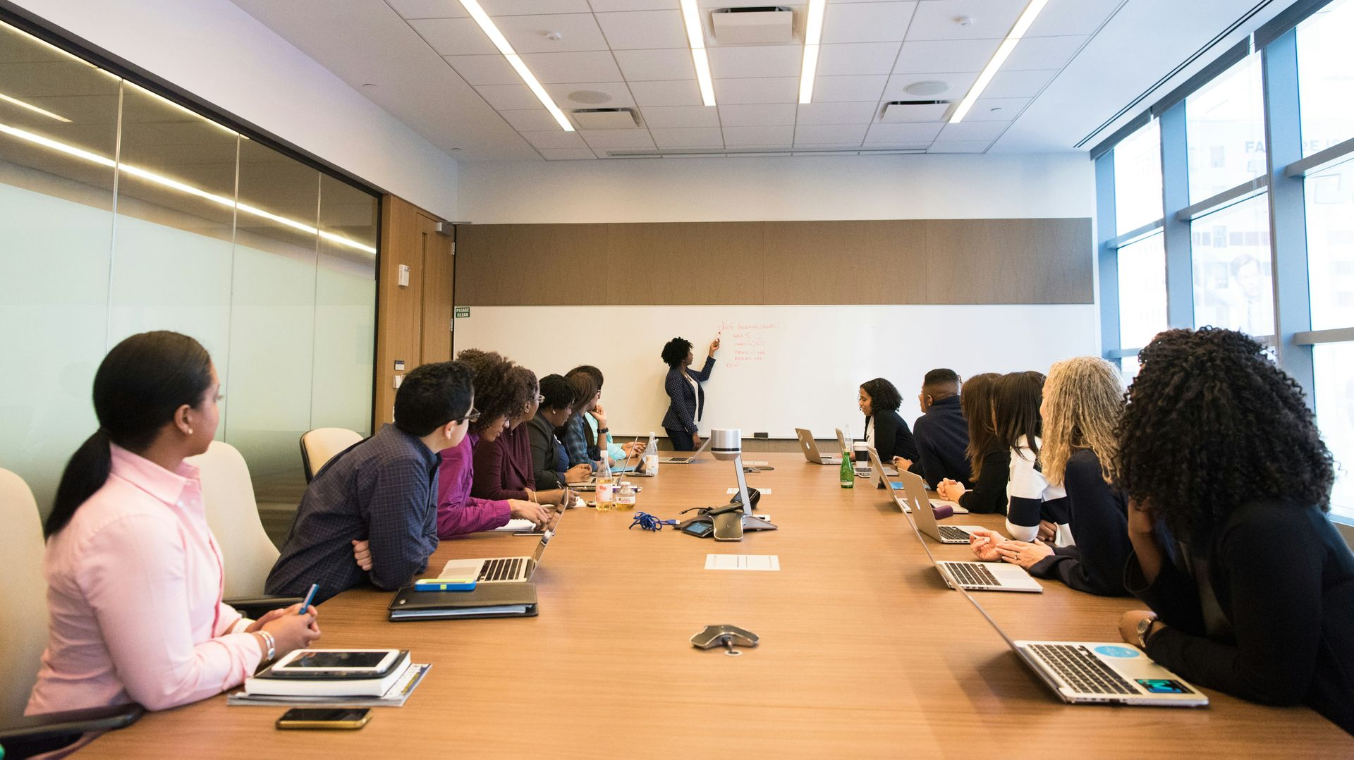People in a conference room, listening to a presentation. A woman writes on a whiteboard.
