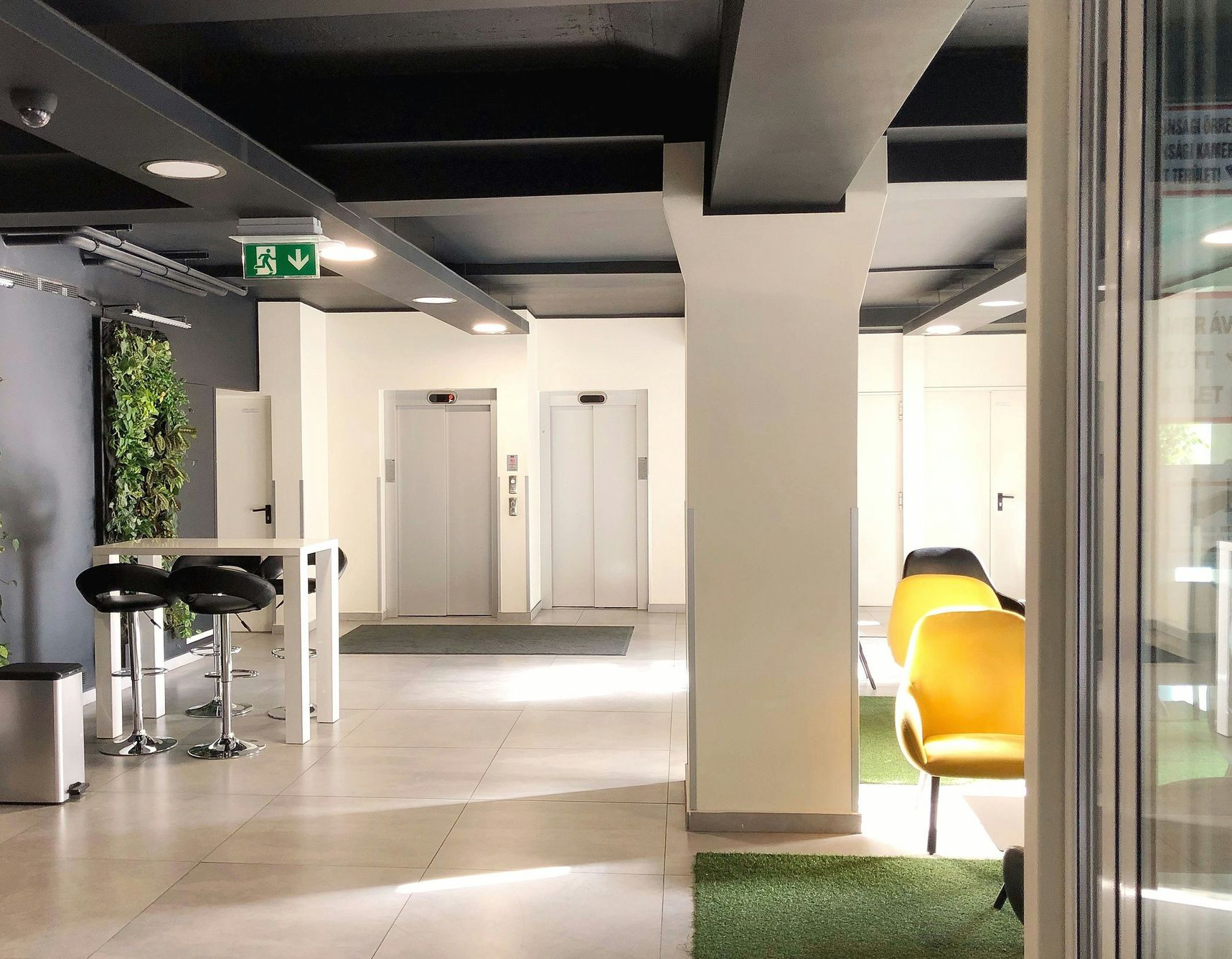 Lobby with two elevators, a bar table with stools, a green wall, and yellow chairs.