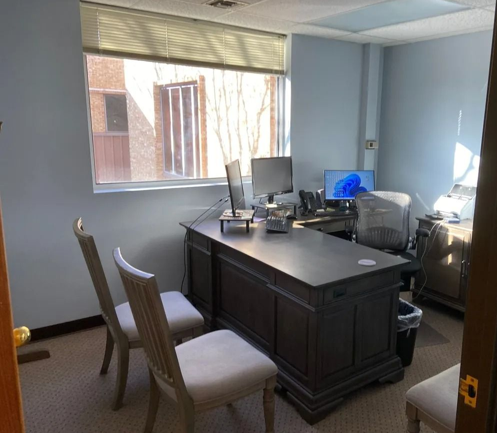Office with a desk, two chairs, and two computer monitors near a window with blinds.