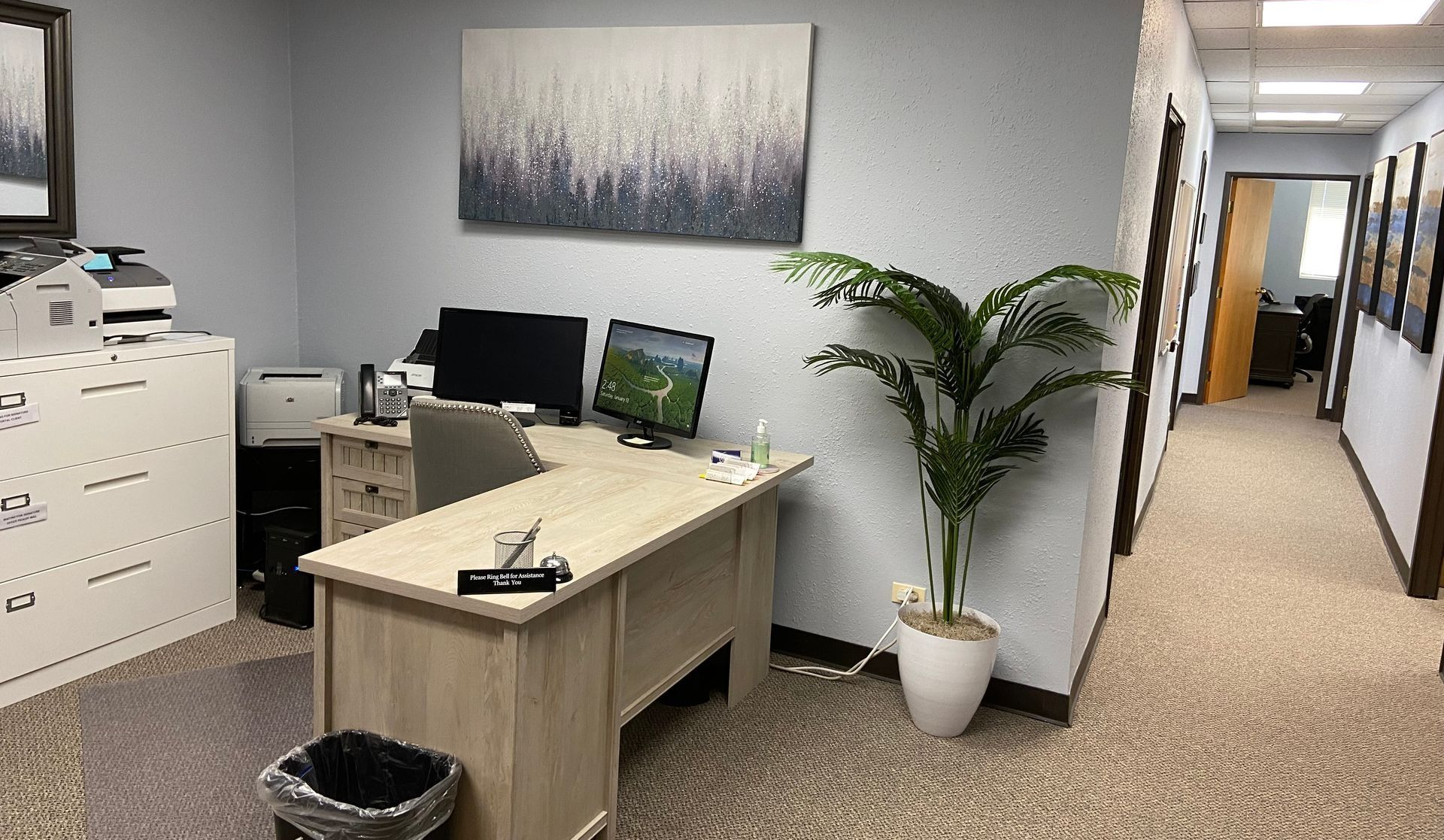 Office with desk, computer, printer, artwork, and a potted plant in a light-blue room, with a hallway visible.