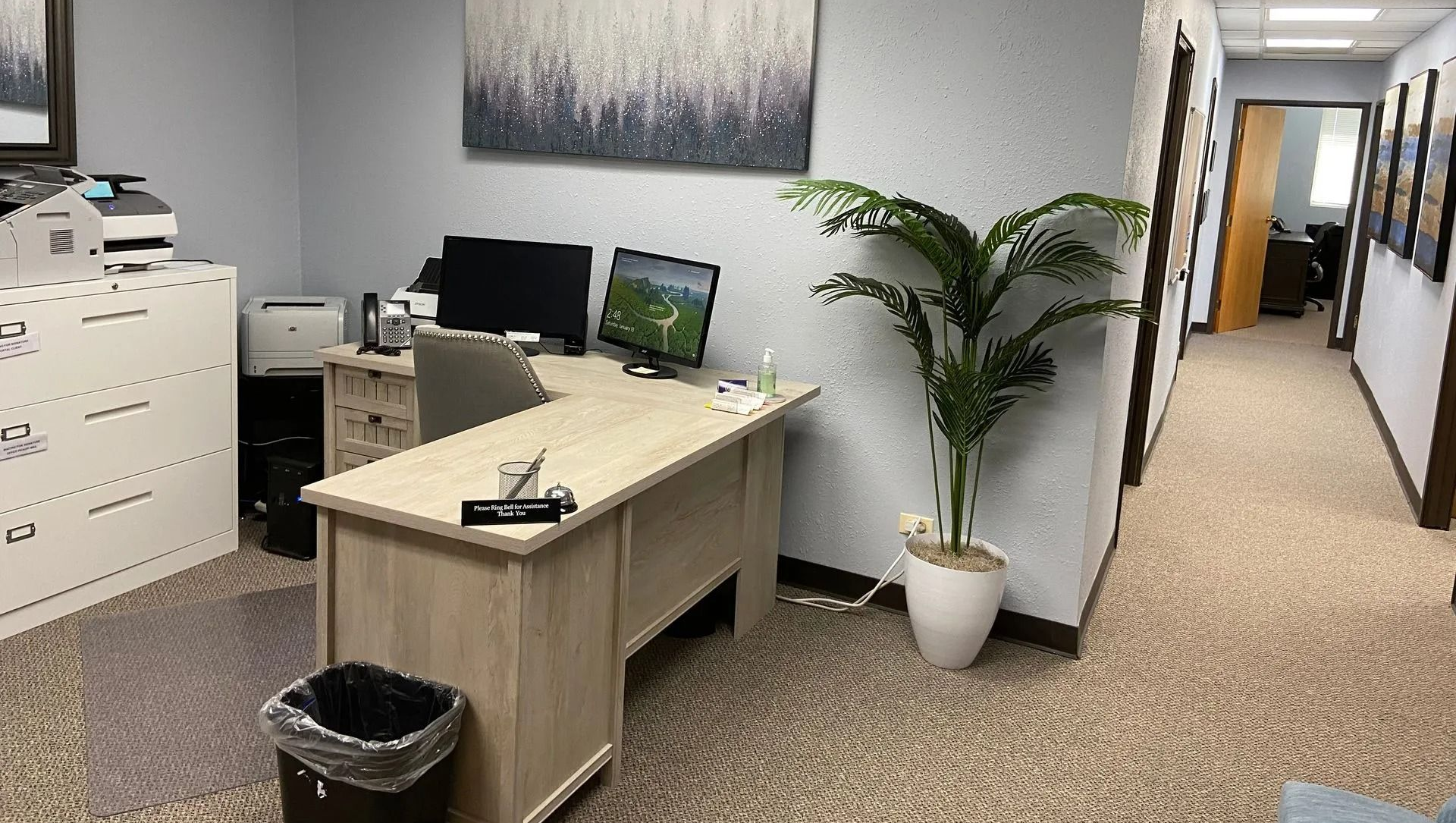 An office with a desk, computers, filing cabinet, artwork, and a hallway.