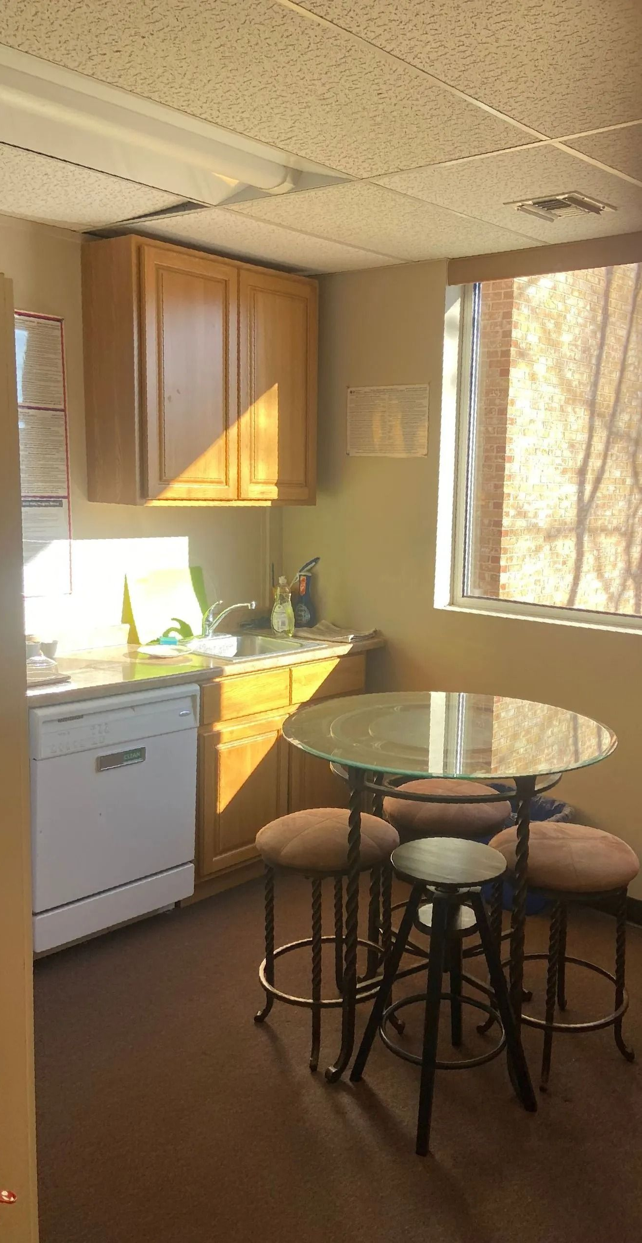A small, sunny kitchen with wood cabinets, white dishwasher, round table, and stools.