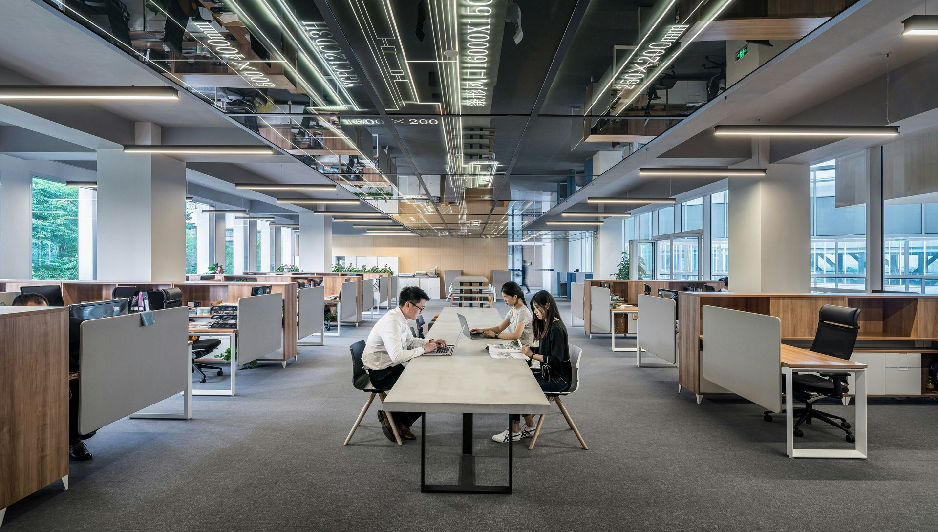 Modern open-plan office with employees working at tables. Light fixtures on mirrored ceiling.