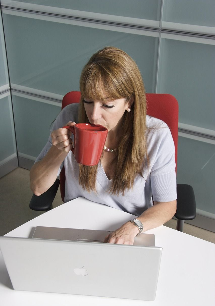 Woman at laptop, drinking from a red mug. Seated in office, typing and drinking.