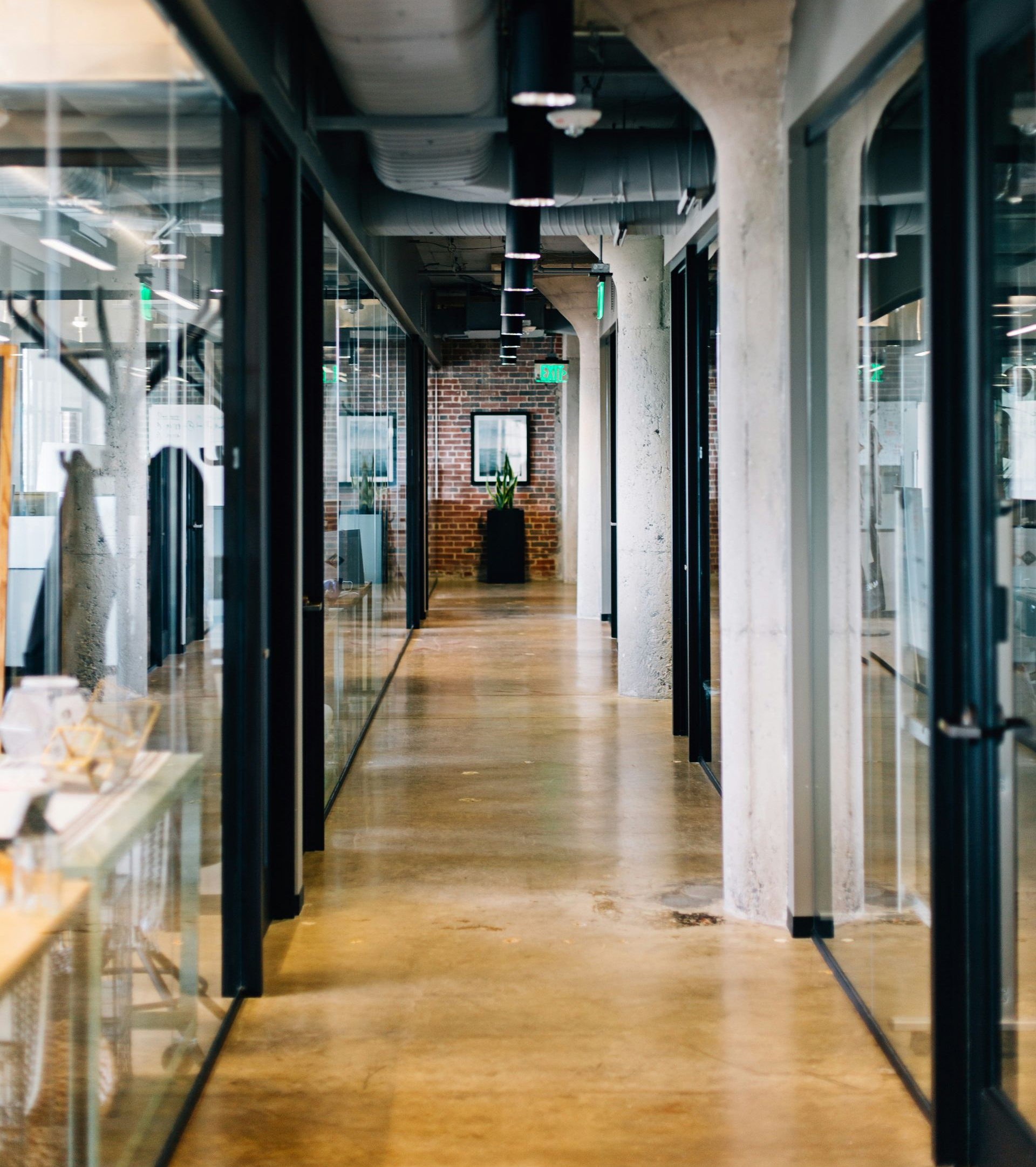 Long, modern office hallway with glass-walled offices, concrete pillars and flooring, and exposed ceiling fixtures.