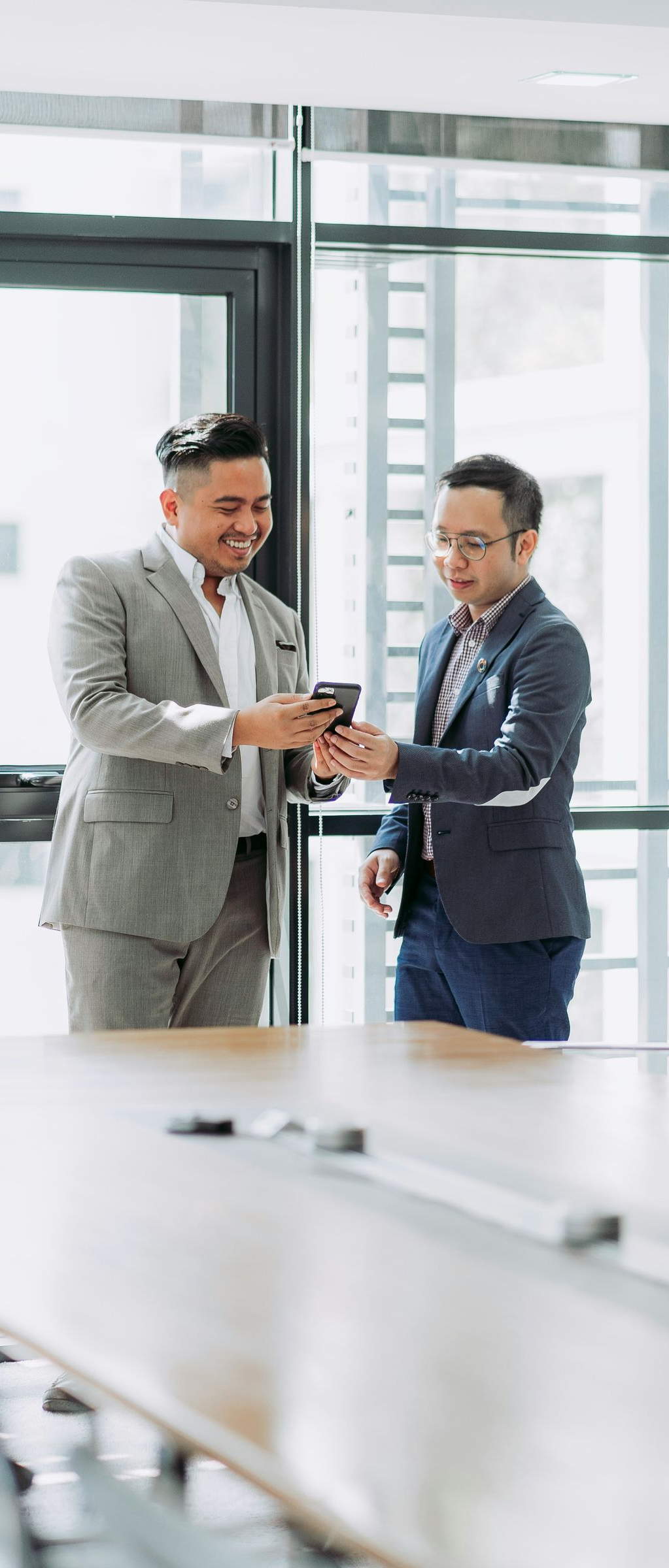 Two men in suits look at a phone in a bright office with a long table.