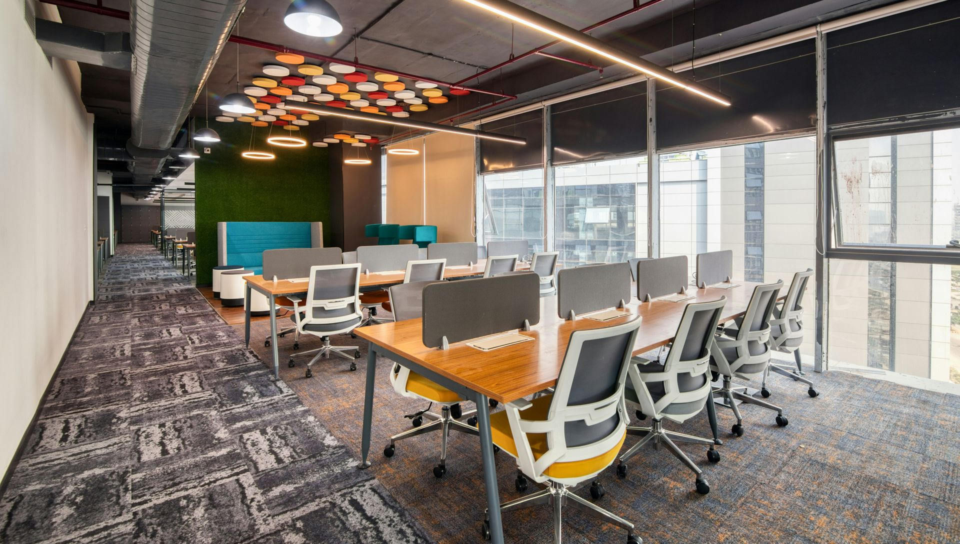 Modern office workspace with desks, chairs, large windows, and decorative ceiling lights.