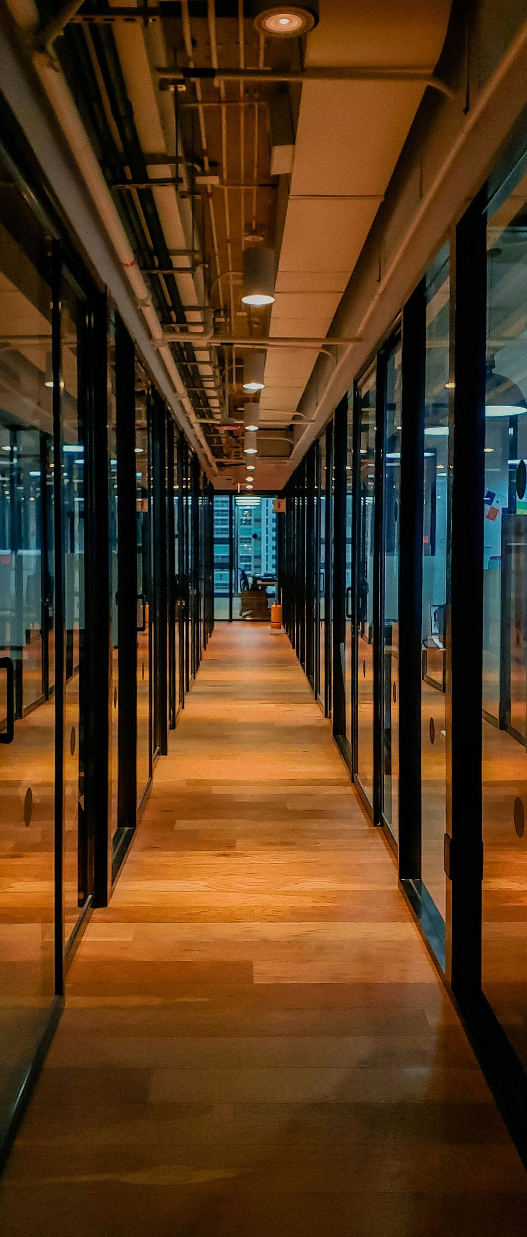 A long office hallway with glass walls, wooden floor, and ceiling lights.