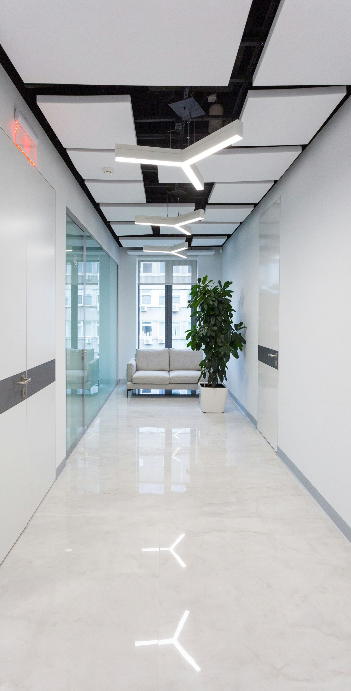 Long hallway with white walls and ceiling lights, ending in a seating area with a plant.