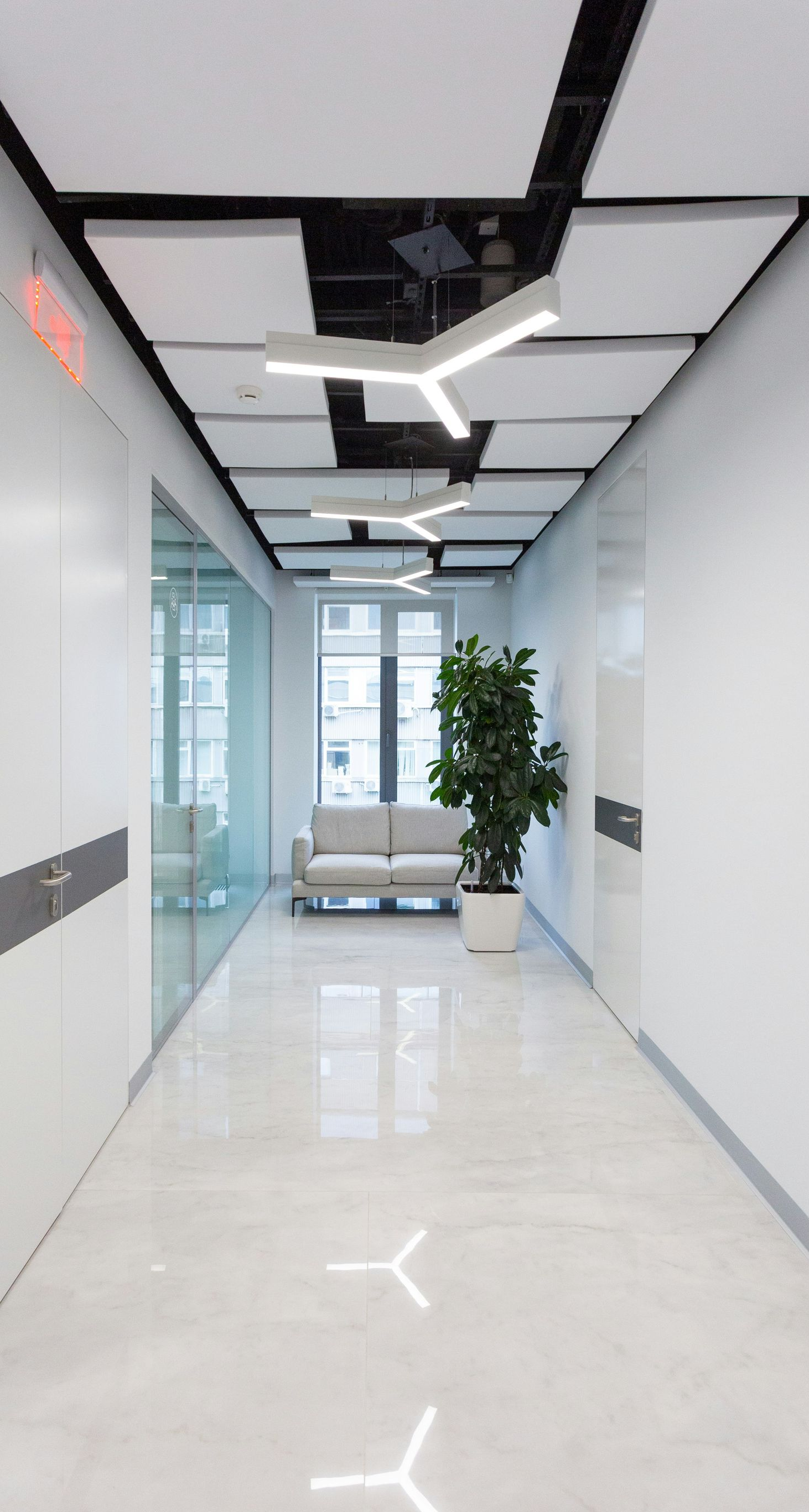 Long hallway with white walls and ceiling lights, ending in a seating area with a plant.