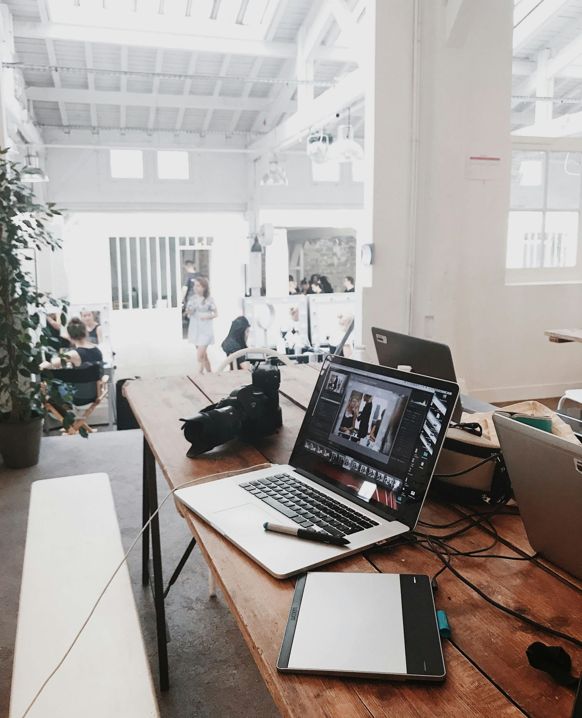 Laptop and camera on wooden table in a bright, open studio space with people in the background.