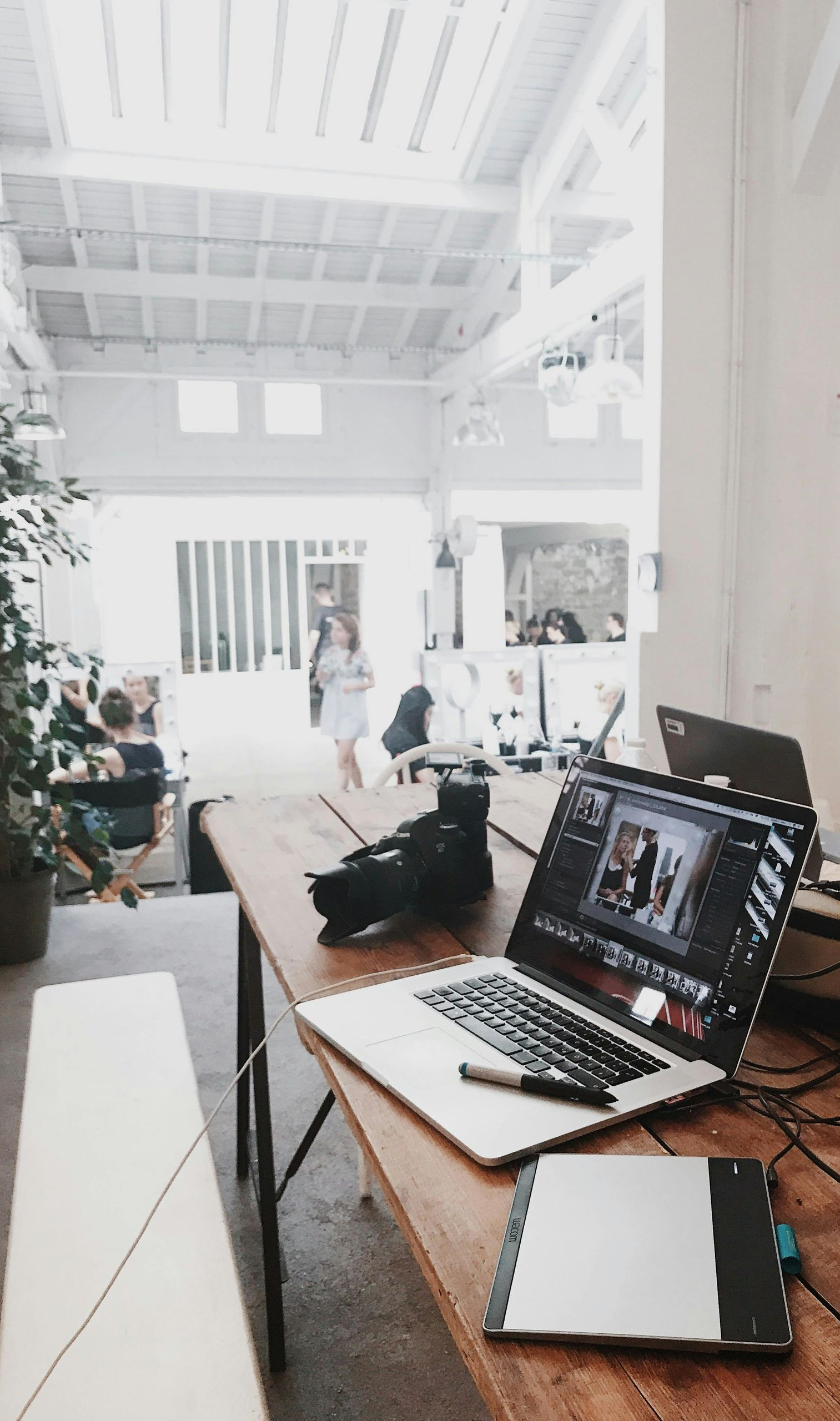 Laptop, camera, and editing tablet on a wood table in a bright, open space. People are visible in the background.