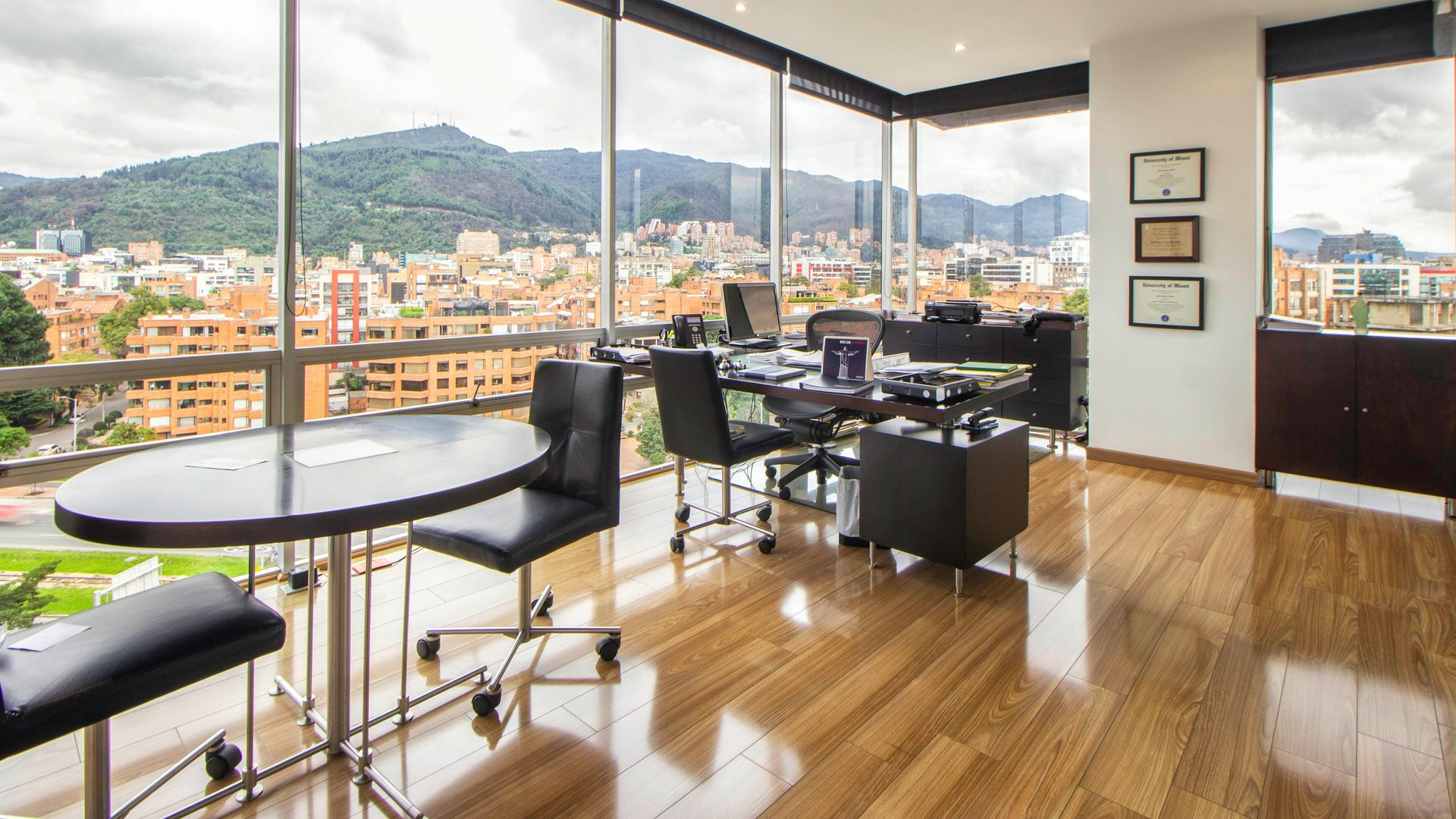 Office interior with large windows, city and mountain view. Table, chairs, desk, and awards on wall.