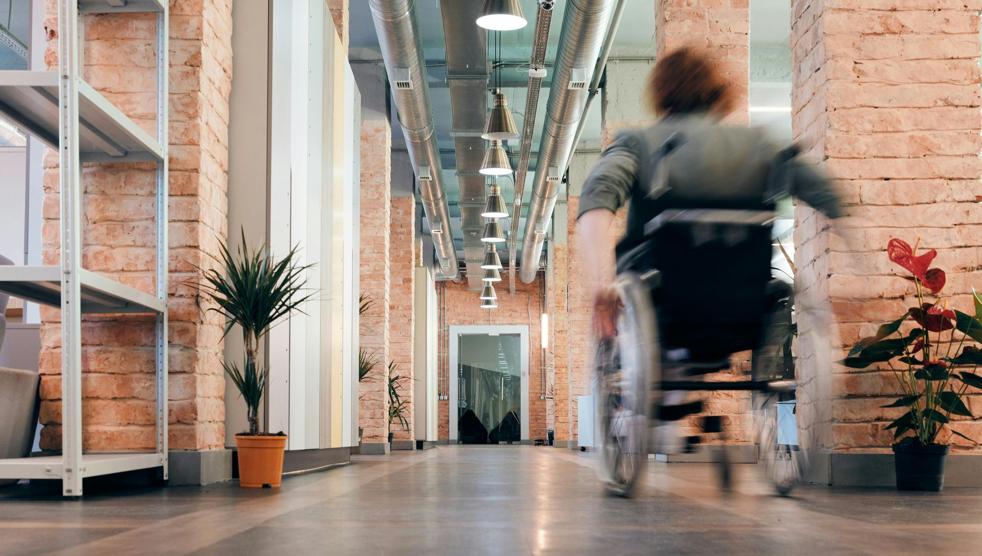 Person in wheelchair moving down a hallway with brick walls and plants.