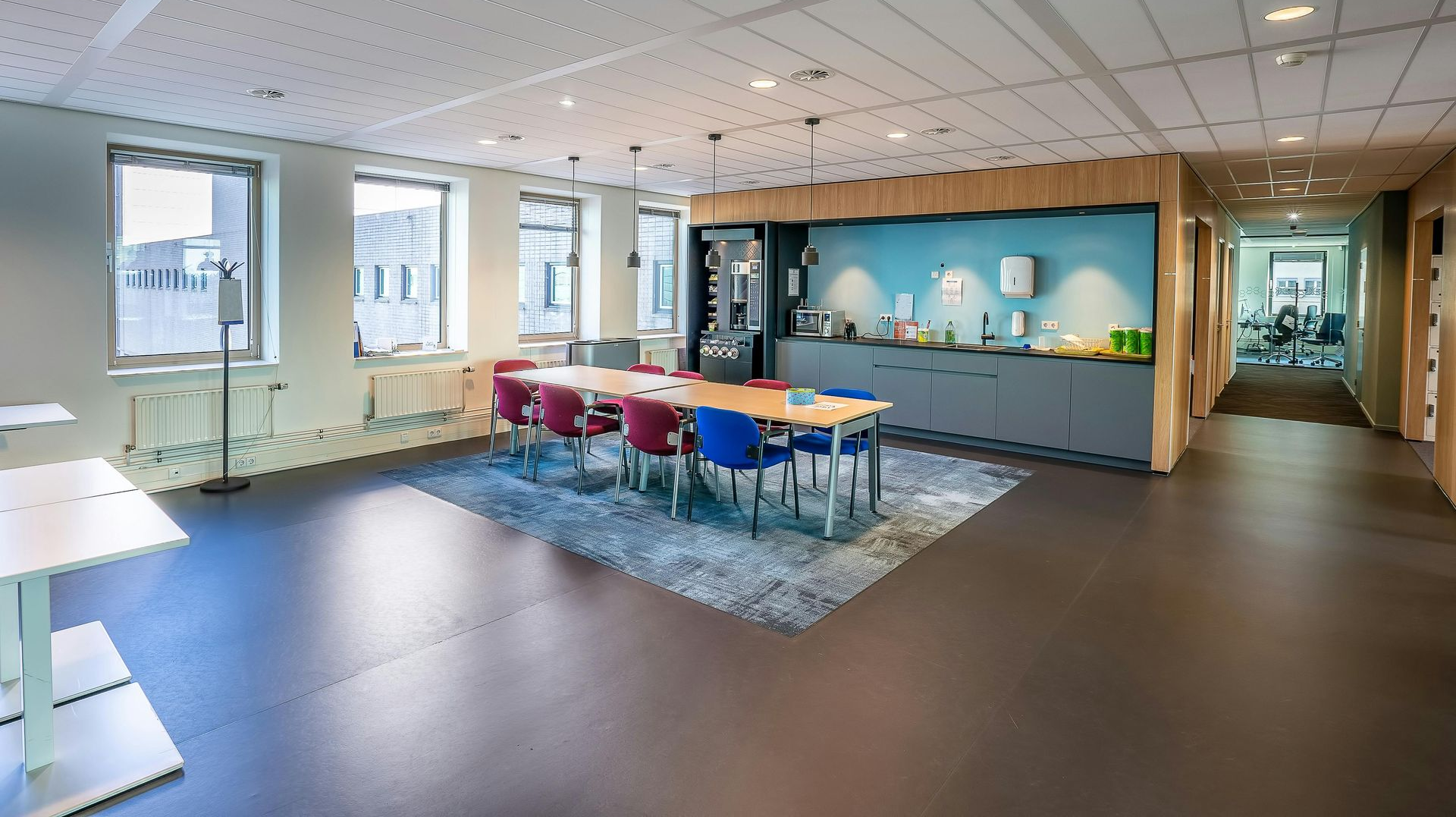 An office breakroom with a long table, chairs, cabinets, and a hallway. Windows on the left.