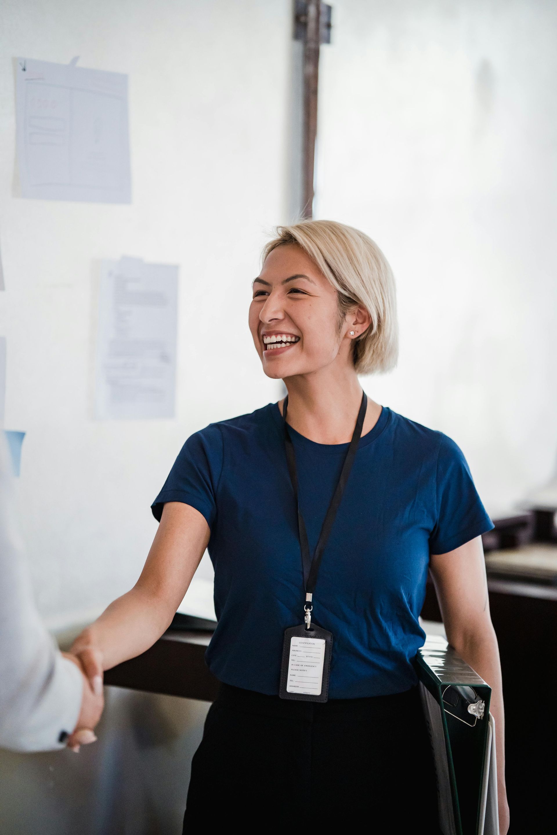 Woman shaking hands, smiling. Wearing blue shirt, lanyard, black pants, in an office setting.