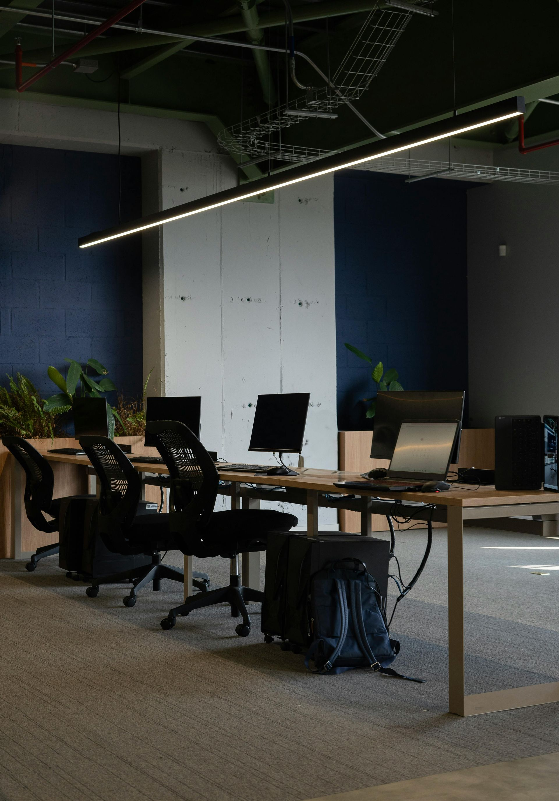 Modern office workspace with desks, chairs, computers, and a backpack; cool tones and overhead lighting.