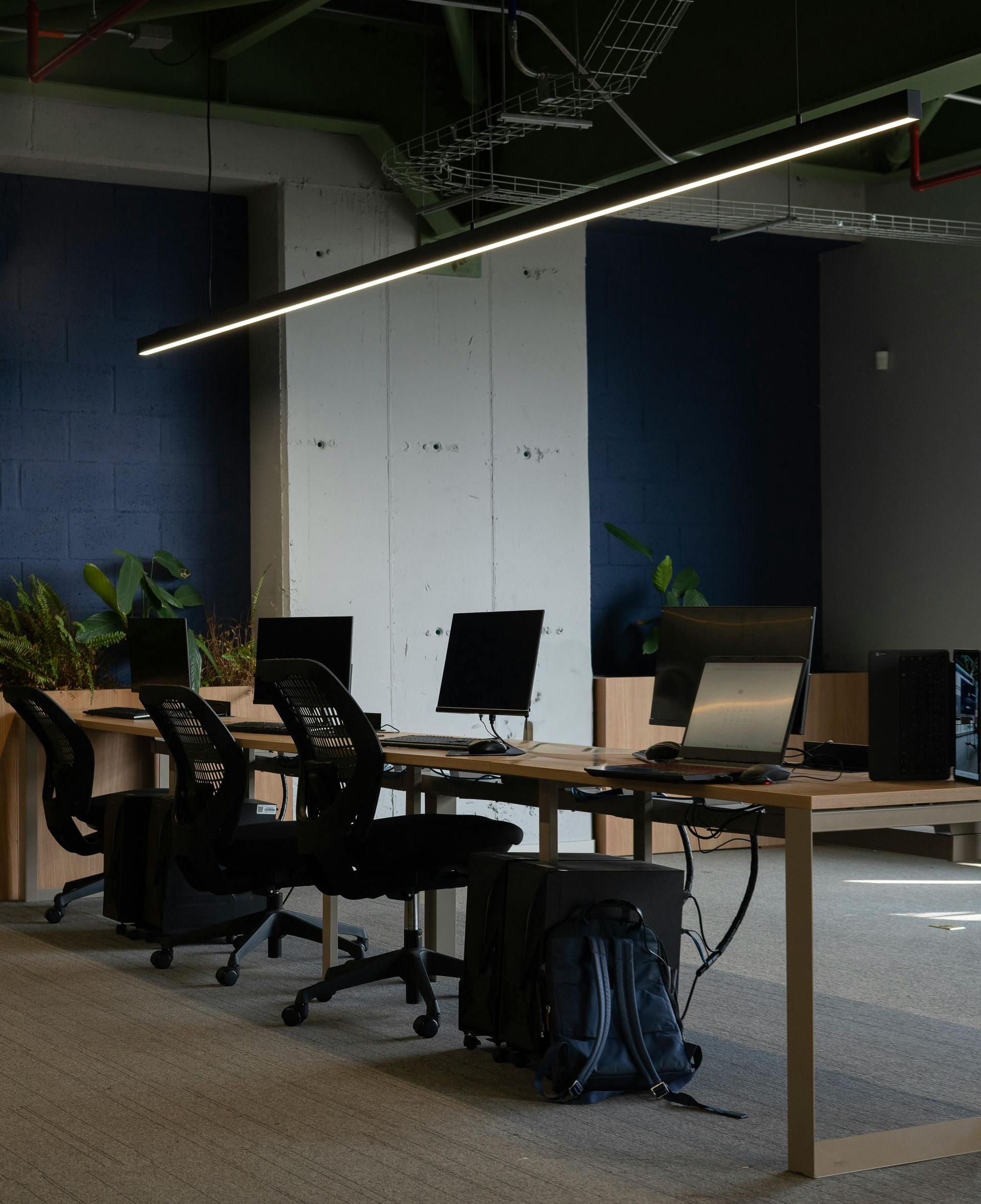 Modern office workspace with desks, chairs, computers, and a backpack; cool tones and overhead lighting.