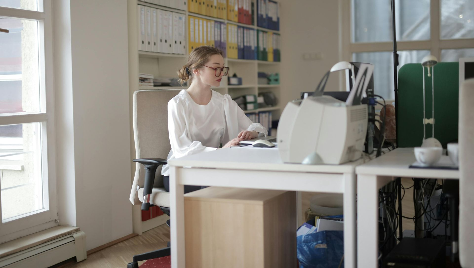 Woman sits at desk in a bright office, working on computer. Shelves with files visible.