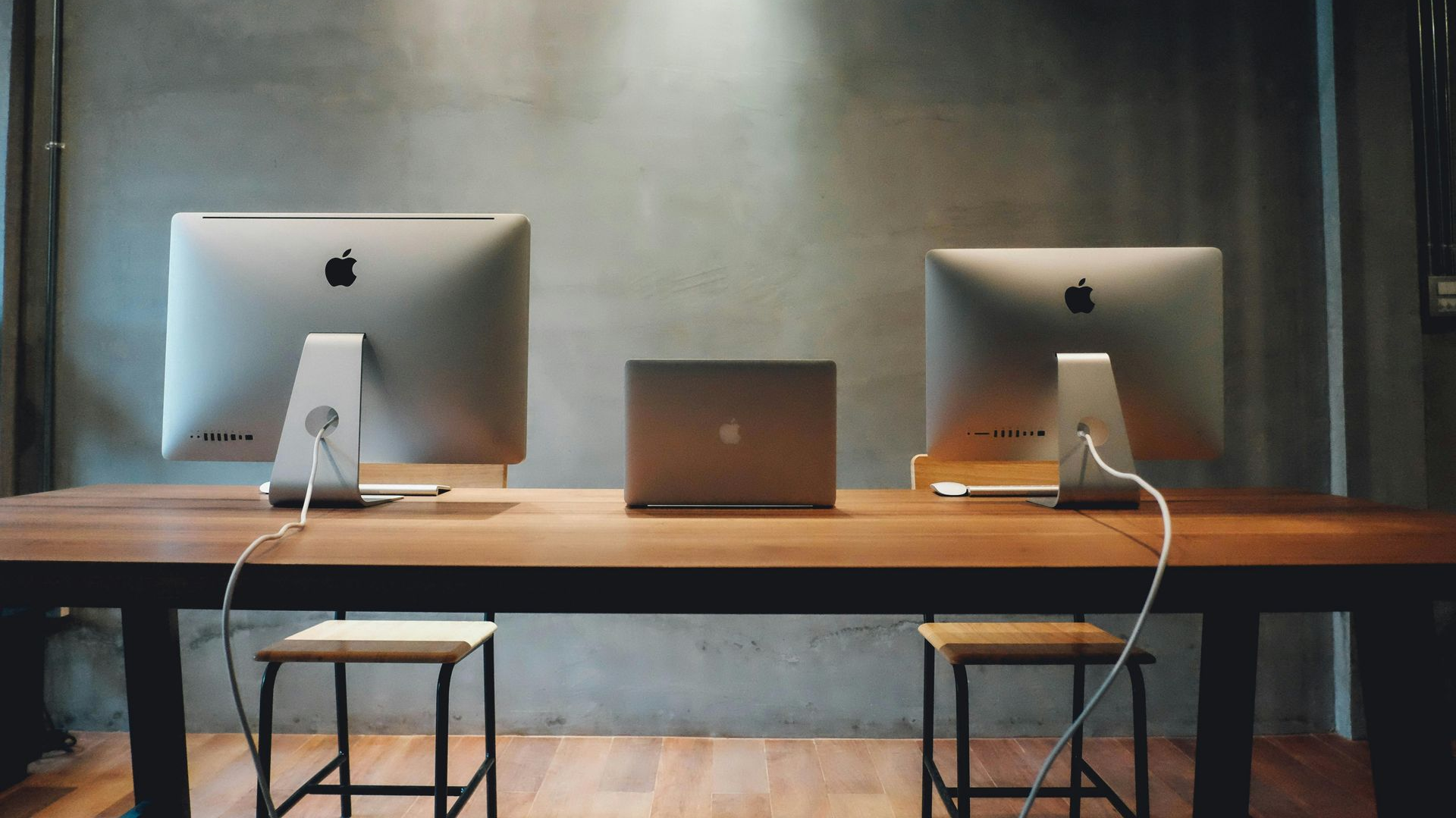 Two iMacs and a MacBook on a wooden desk with two stools in front of a gray wall.