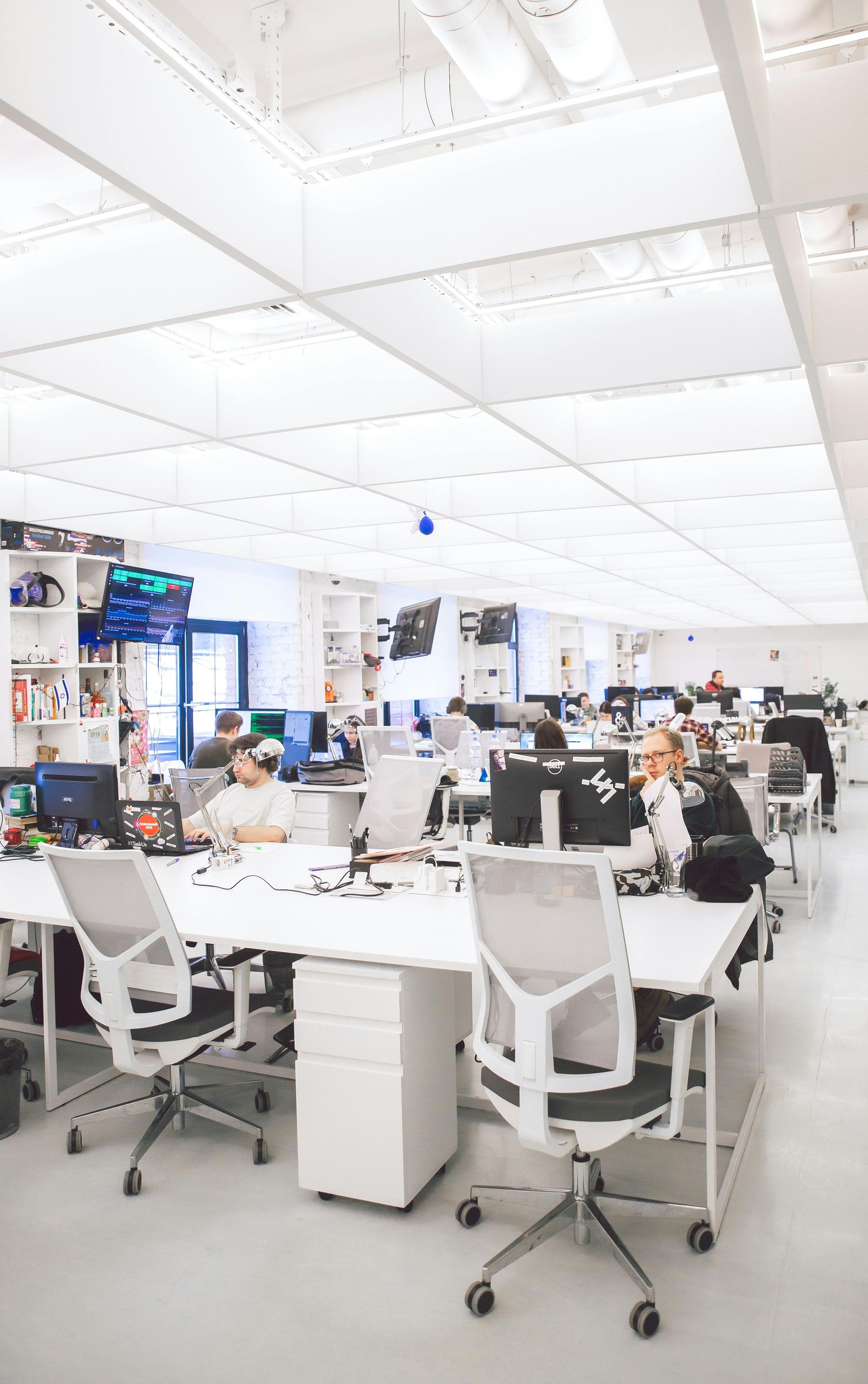 Bright, modern office space with white desks and chairs. People work at computers. White ceiling with recessed lighting.