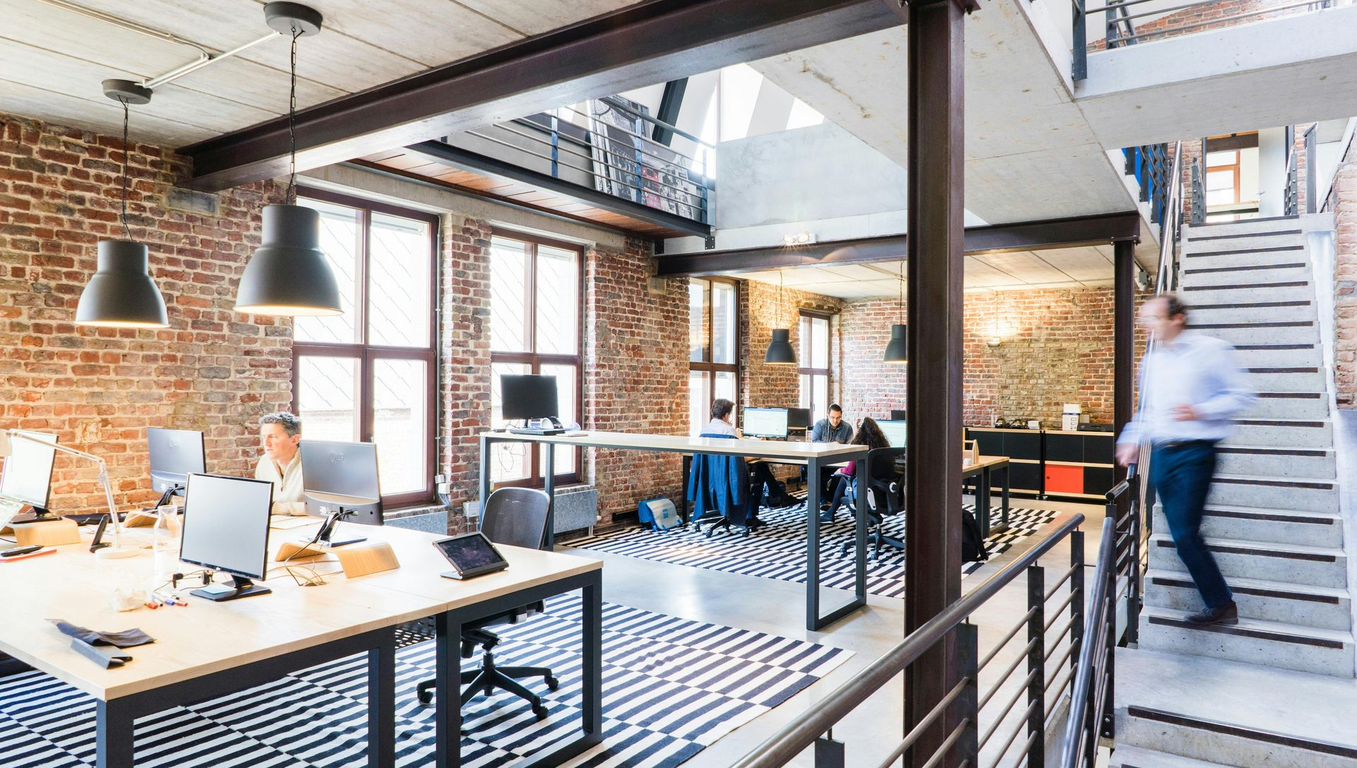 Modern office with desks, exposed brick, and a person walking down stairs.