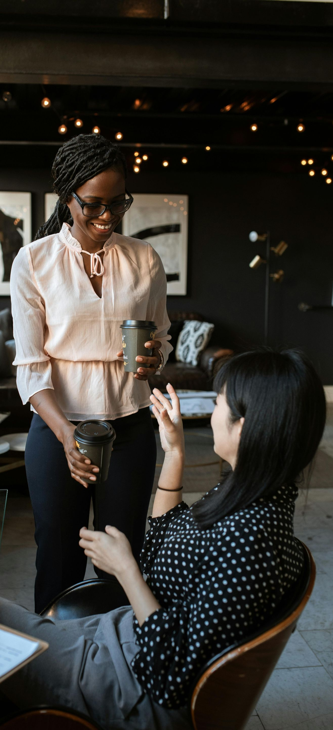 Two women talking at a table. One holds a glass and a coffee cup, and the other gestures while seated.