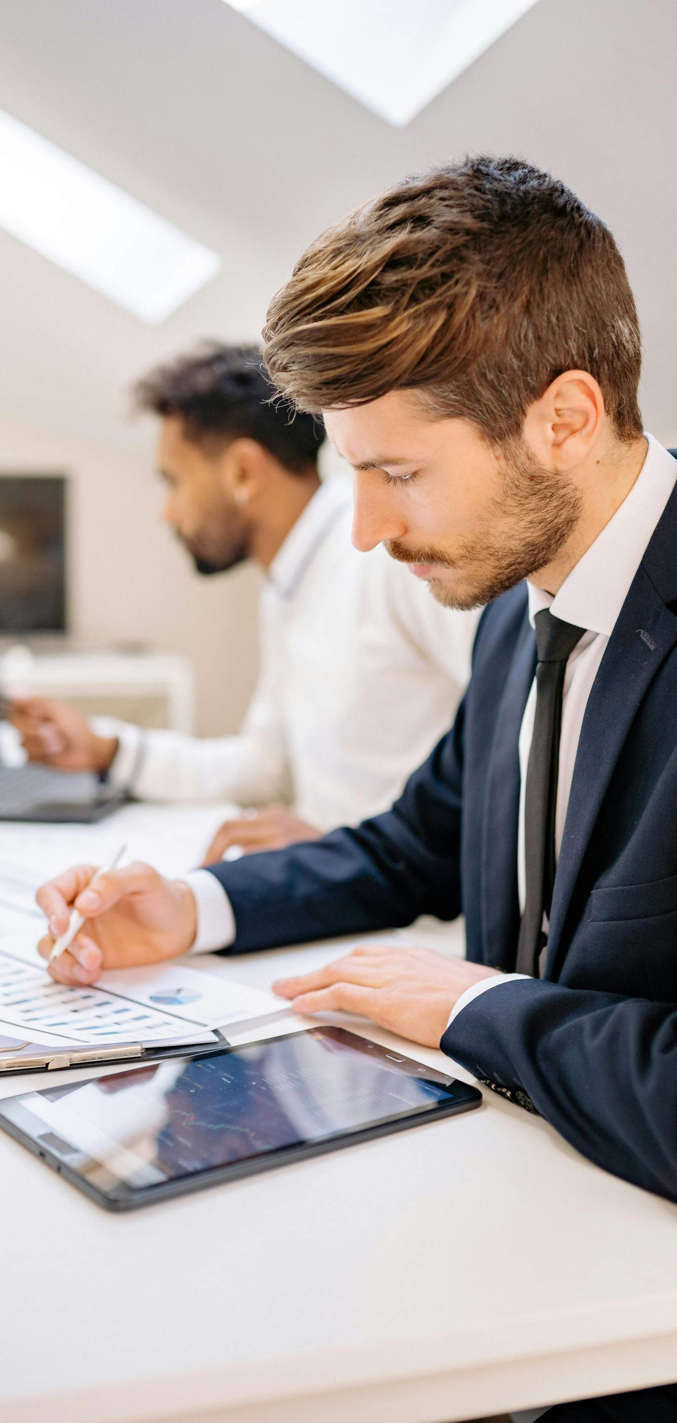 Man in suit working, with a colleague, at a desk with papers and a tablet.