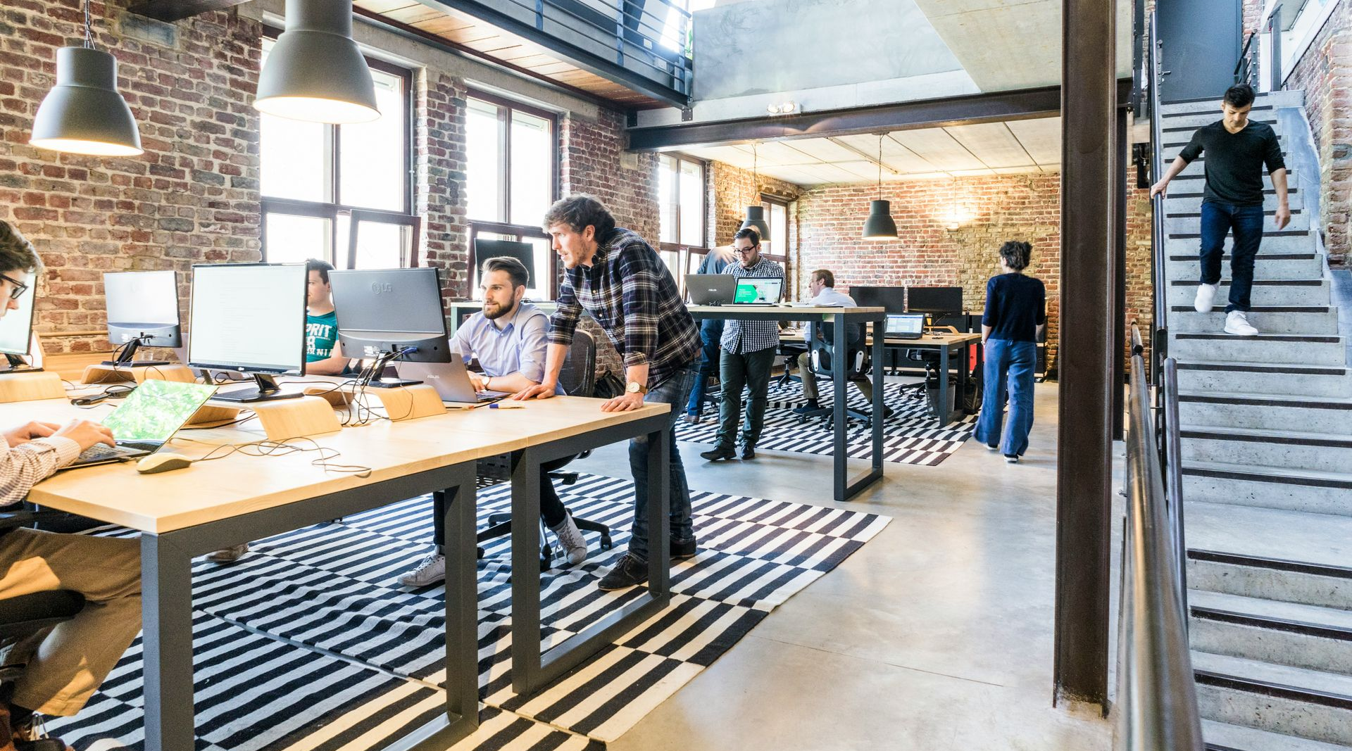 Open-plan office with people working at desks and on stairs, exposed brick, black and white rug, overhead lights.