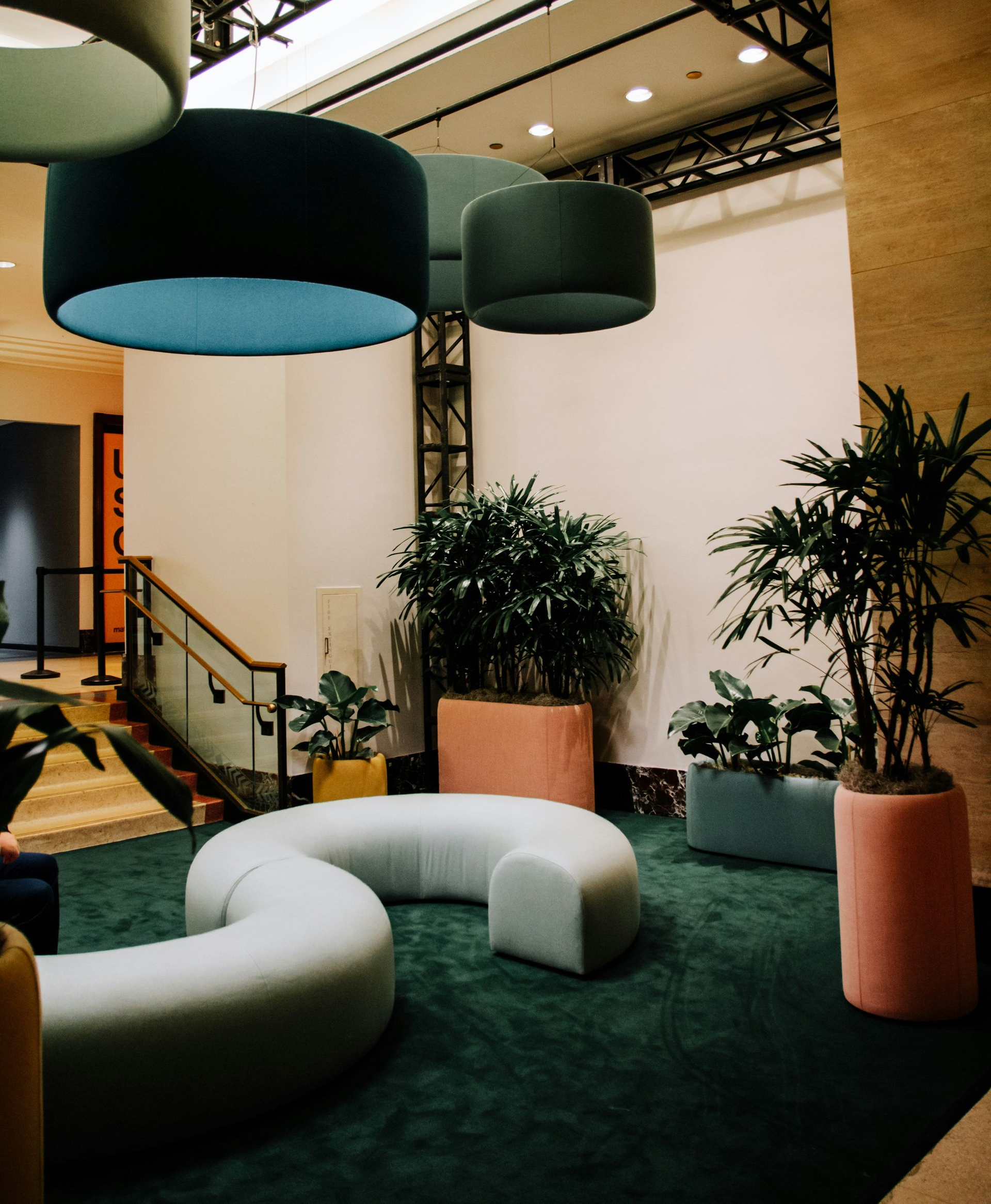 Lounge area with teal pendant lights, curved white sofa, and colorful planters with plants, in an office space.
