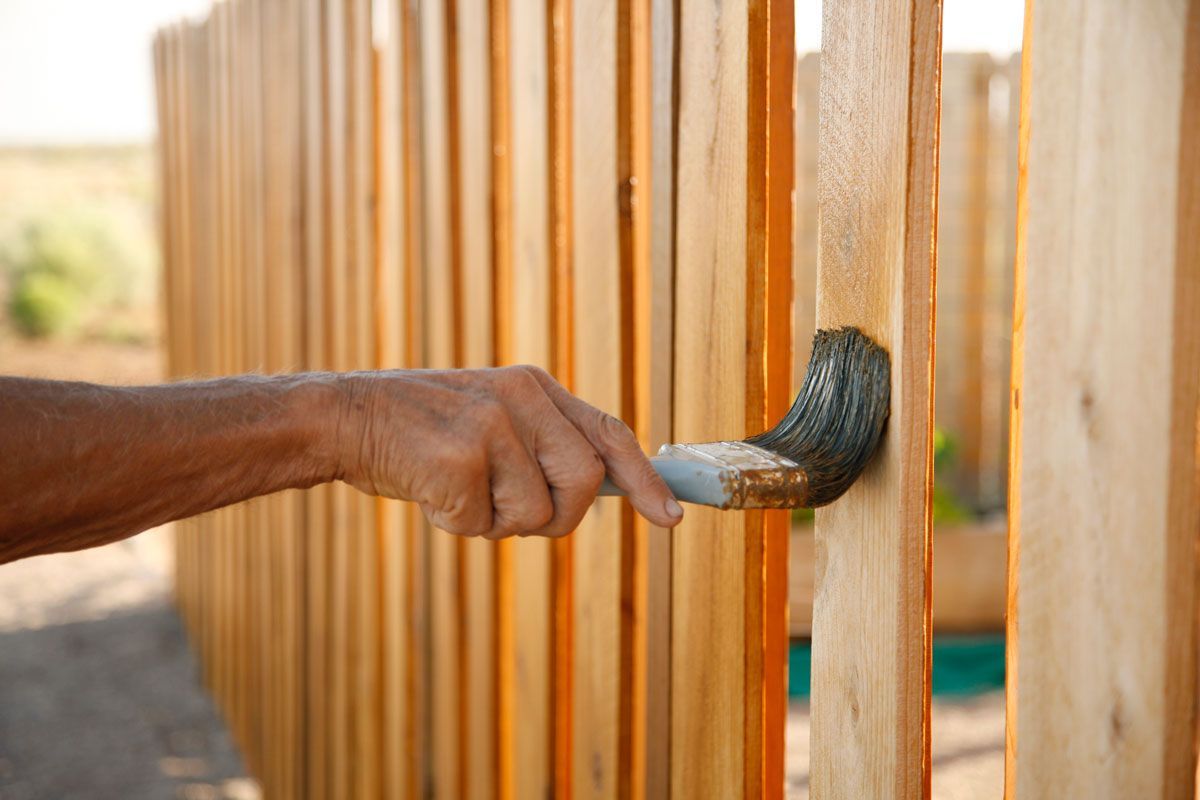 Person staining a wooden fence with a brush outdoors.