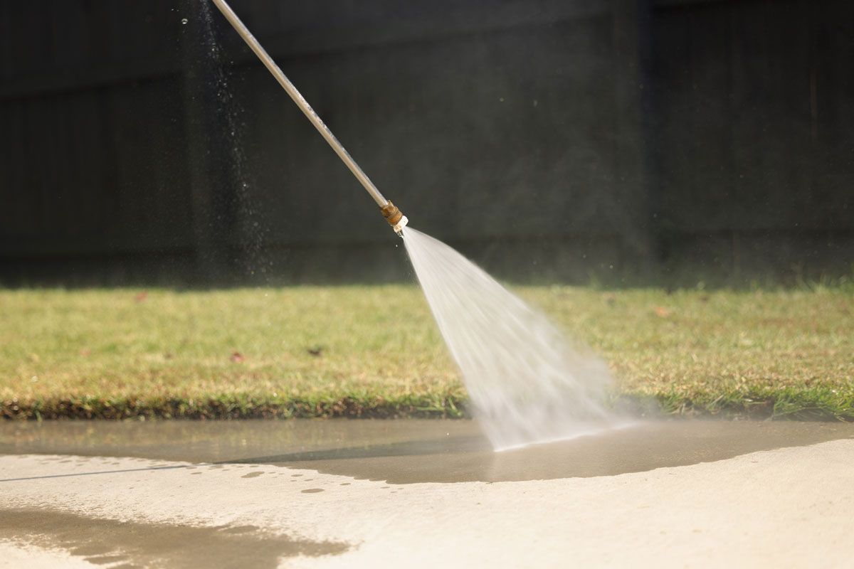 A pressure washer sprays water onto a concrete surface, creating a fan-shaped spray and mist in a yard.