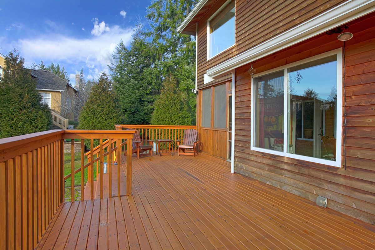 Wooden deck beside a brown house, two chairs, and view of a lush green yard under a partly cloudy blue sky.