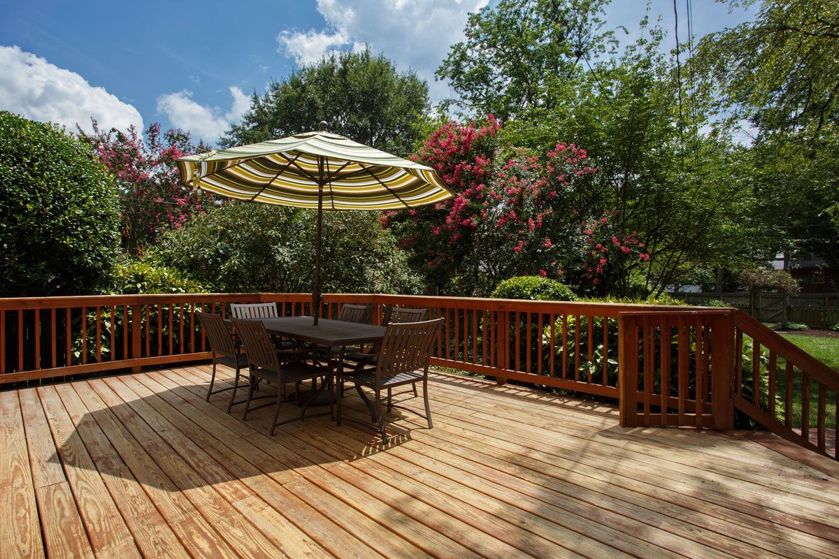 Wooden deck with table, chairs, and umbrella in a sunny backyard surrounded by trees and bushes.