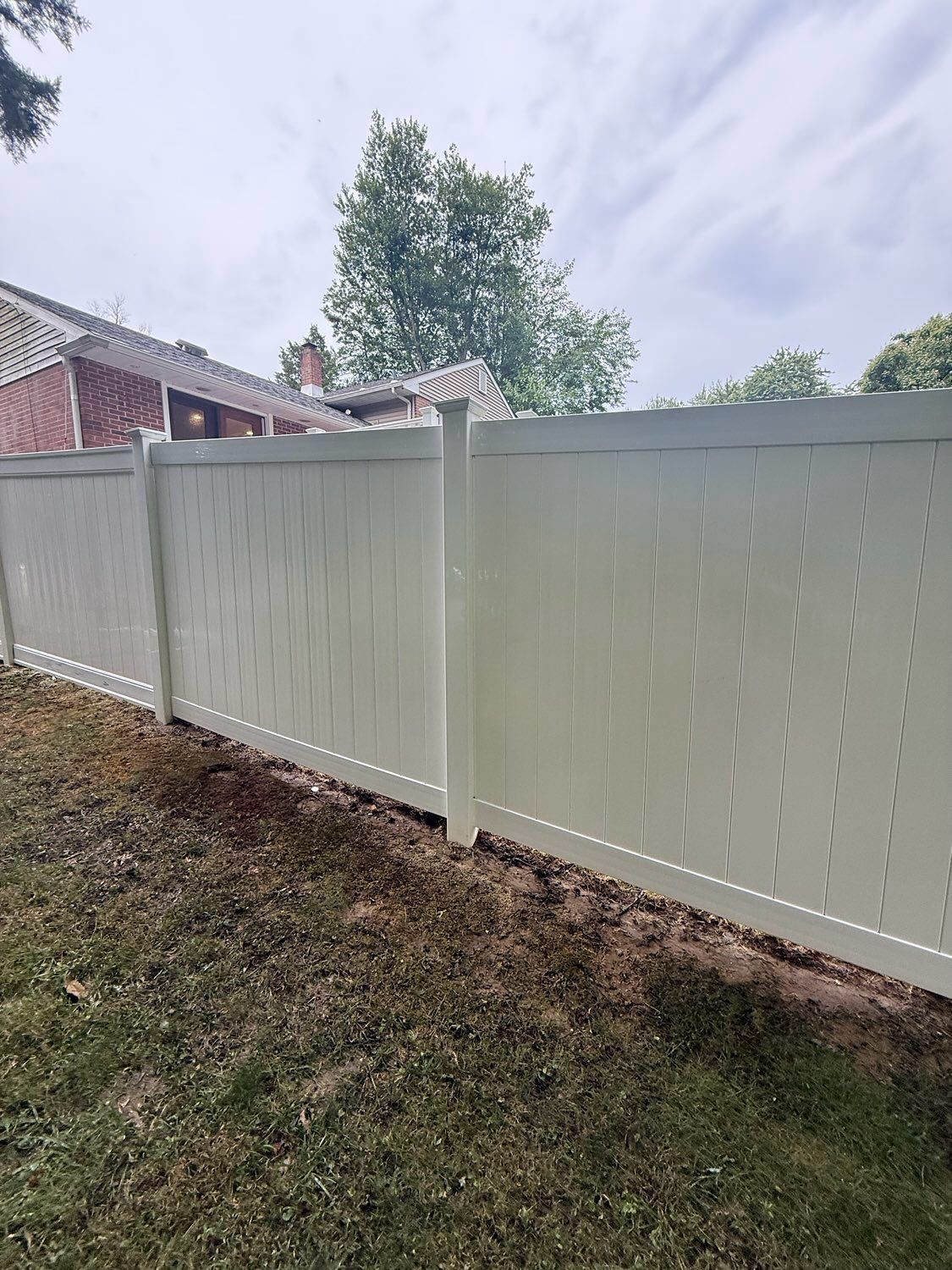 A white vinyl privacy fence stands in a backyard with a house visible in the background under a cloudy sky.