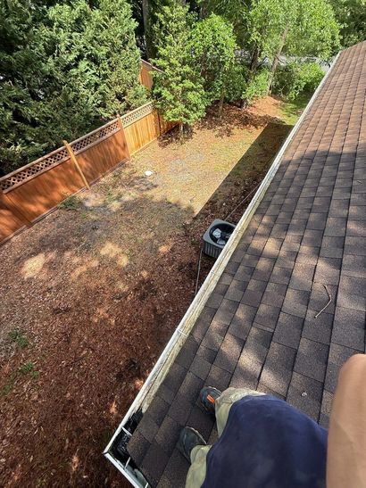 High-angle view from a brown shingled roof looking down onto a mulch-covered yard, wooden fence, and surrounding trees.