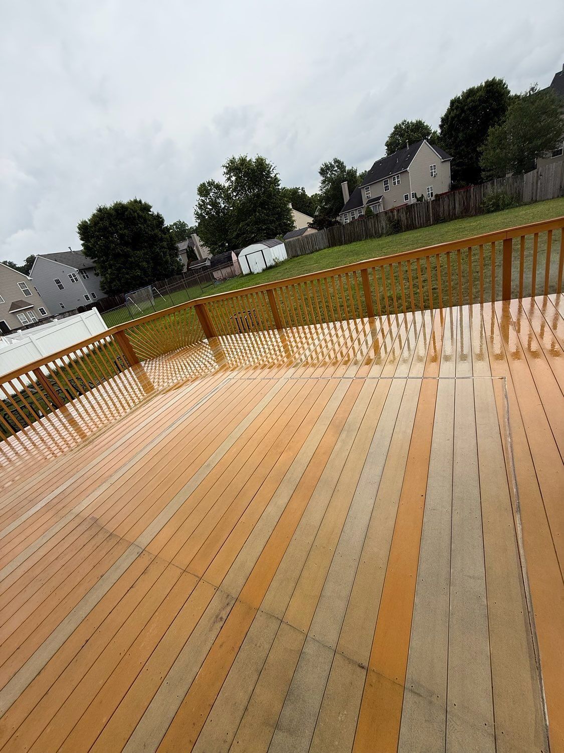 A view of a large, light-brown wooden deck with a matching railing, overlooking a grassy backyard and neighboring houses.
