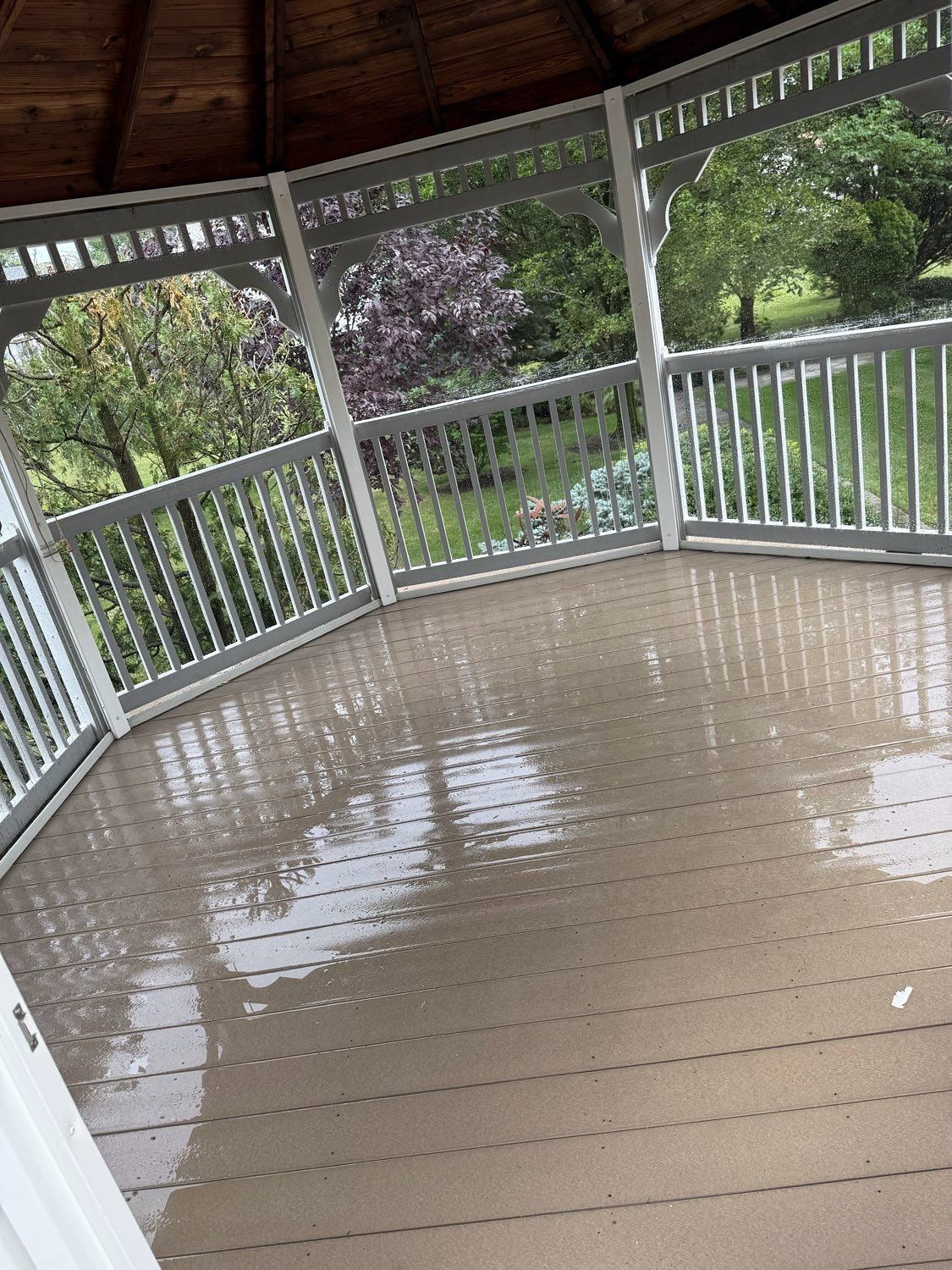 A wooden gazebo with a wet floor reflecting trees and greenery on a rainy day.