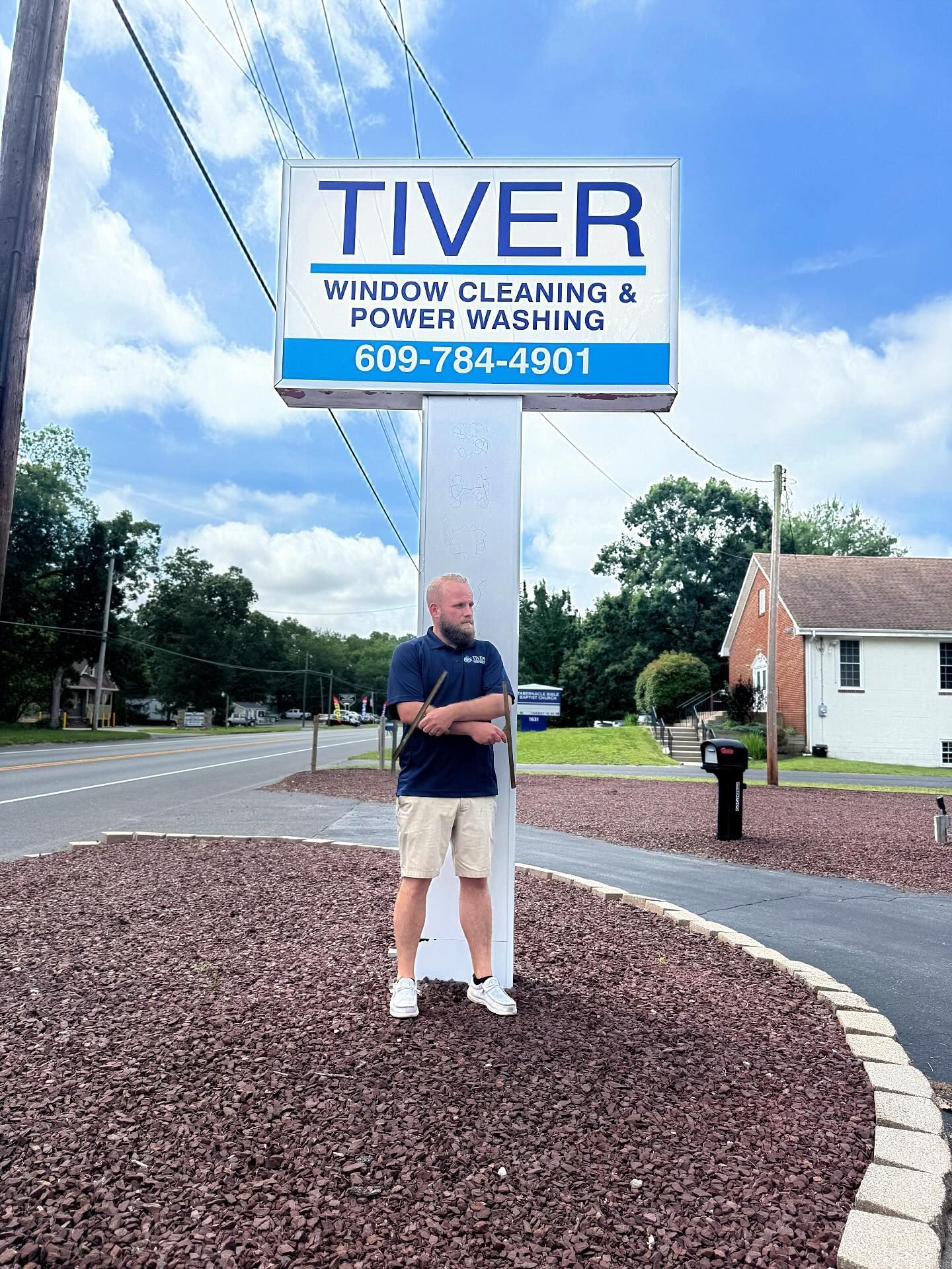 A person stands with arms crossed in front of a Tiver Window Cleaning & Power Washing sign on a sunny day.