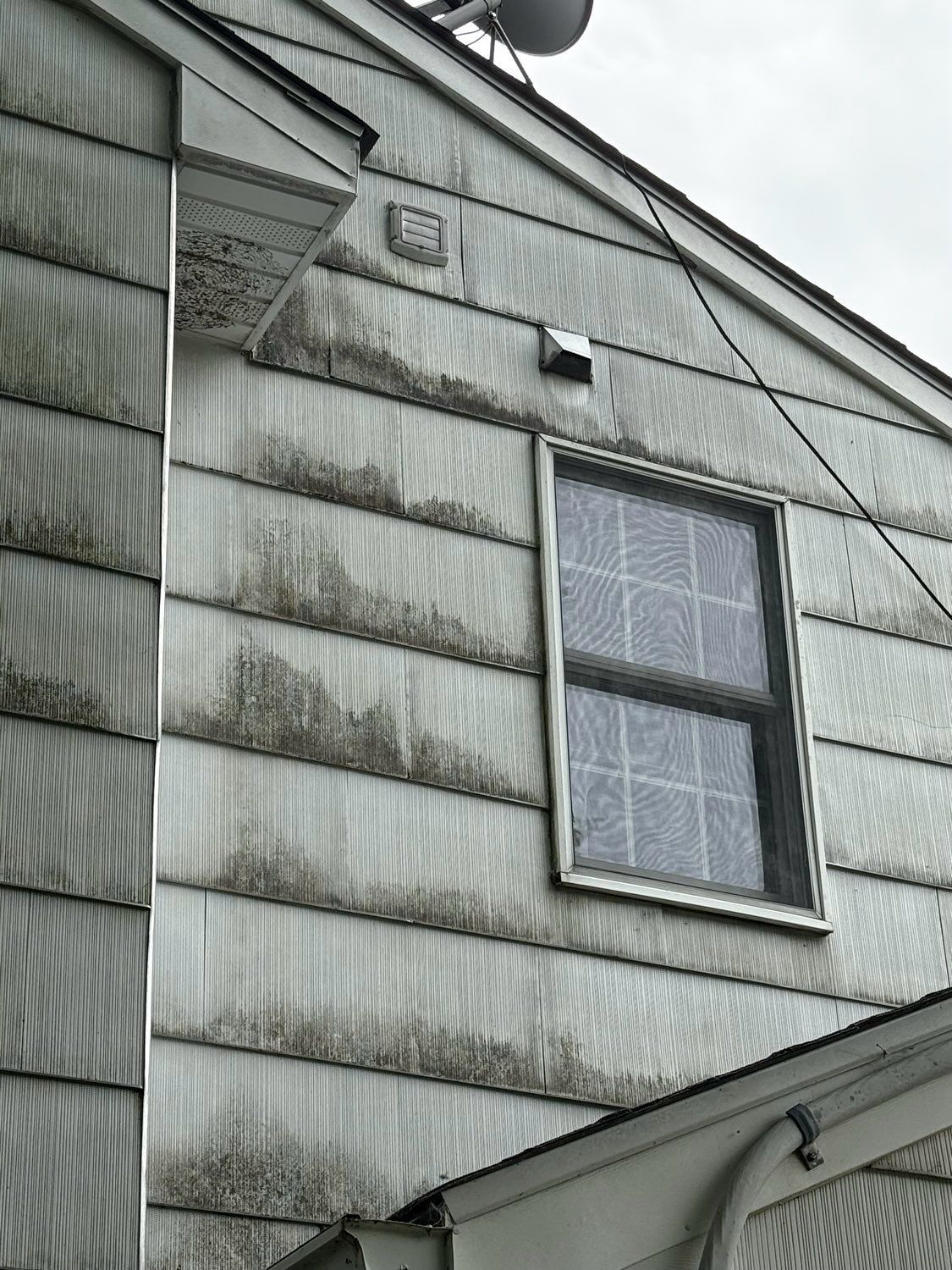 A low-angle view of a grey house exterior with stained, weathered siding and a single window under a cloudy sky.