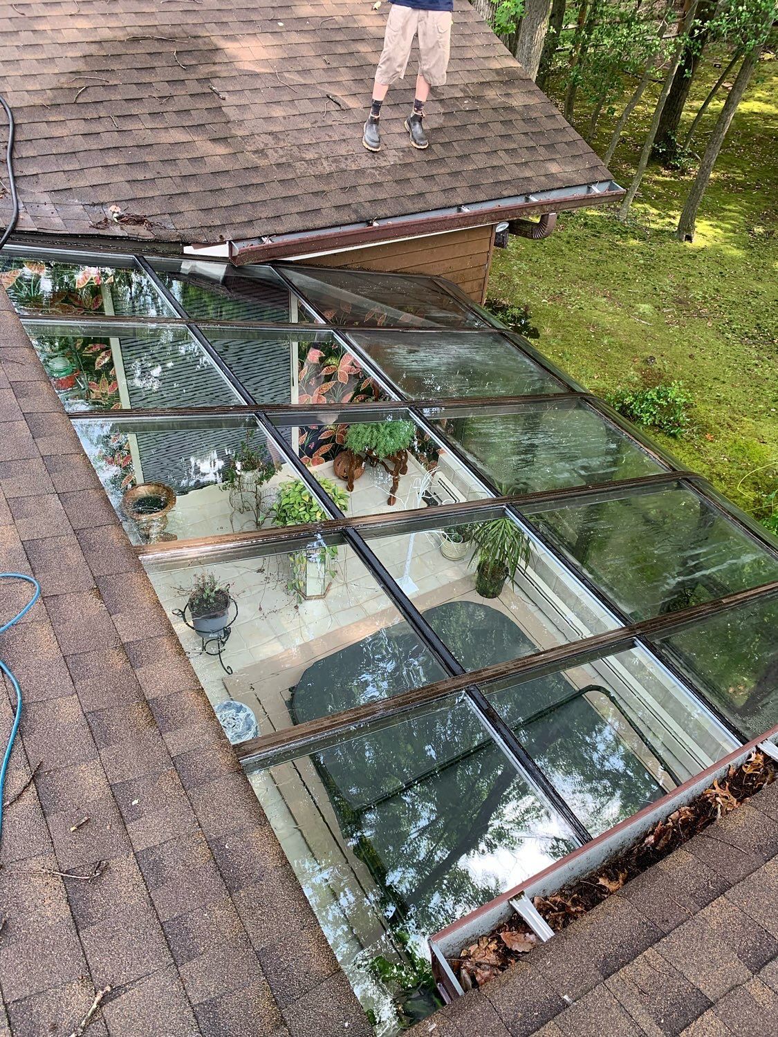A person stands on a shingled roof looking down through a large, rectangular glass skylight at a room filled with plants.