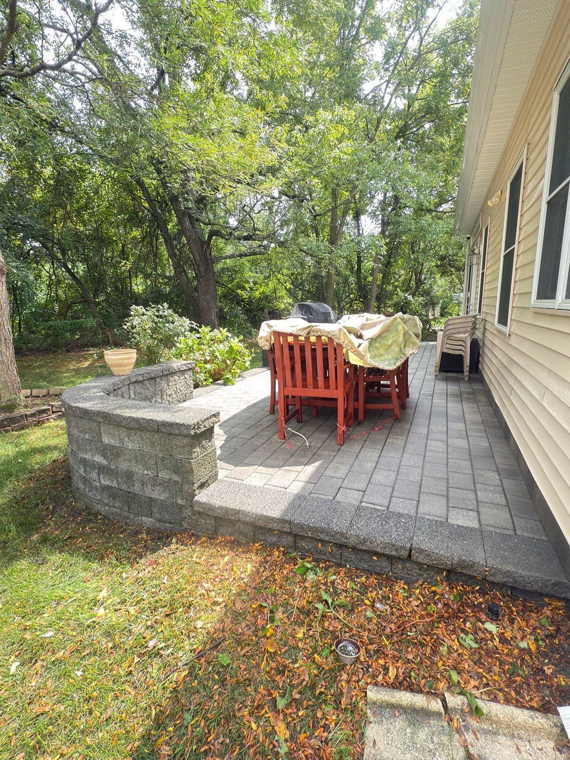 A paved patio with red wooden chairs and a table covered by a light-colored cloth, set against the side of a house.