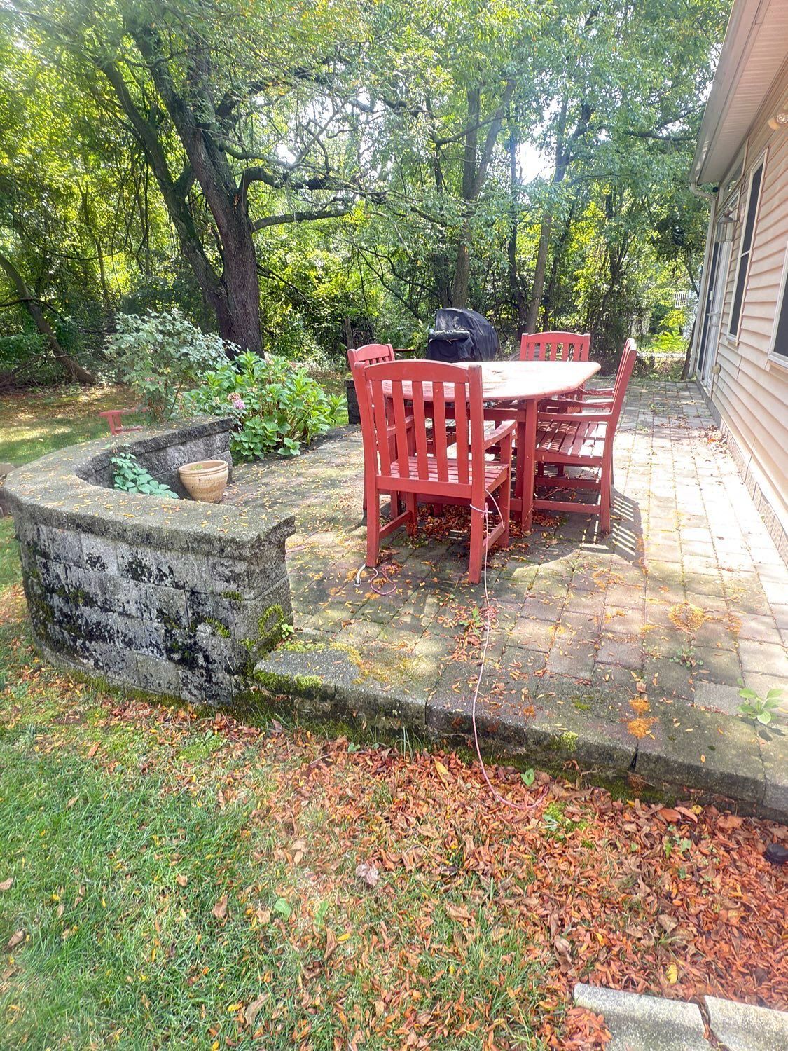 A patio with a round white table and red wooden chairs set against a stone retaining wall and lush green trees.