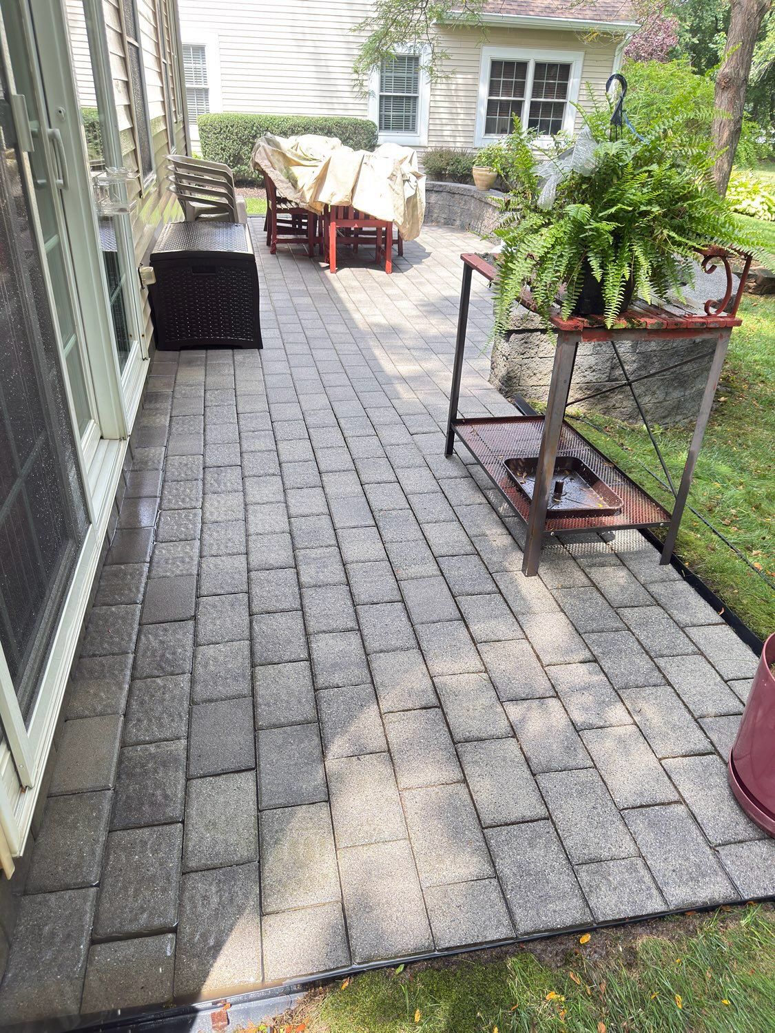 A stone patio with a metal plant stand, a covered outdoor table and chairs, and a sliding glass door at the side.