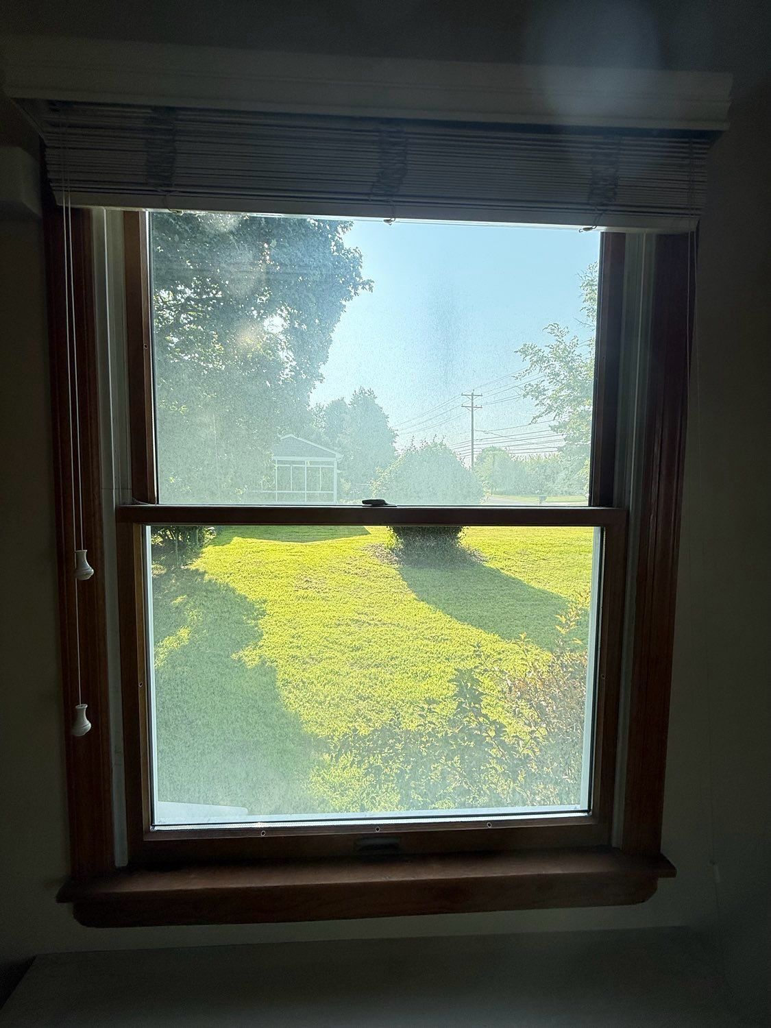 A rectangular window with dark wood trim and a textured shade, looking out onto a bright, grassy yard with trees.