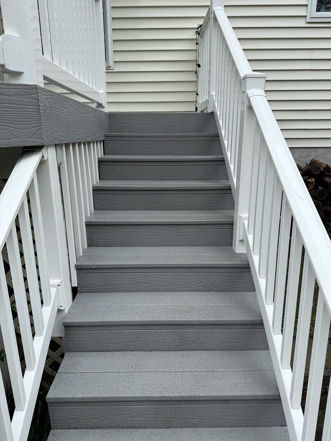 A straight set of grey wooden outdoor stairs with white railings leading up to the side of a house with light siding.